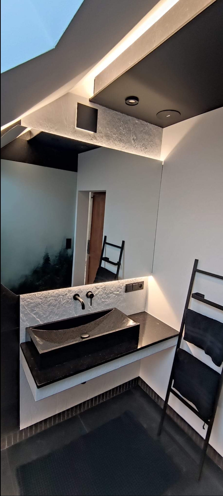 Nowoczesna łazienka z czarną umywalką nablatową o nieregularnym kształcie, czarną armaturą i podświetlanym lustrem, uzupełniona o dekoracyjny grzejnik drabinkowy z ręcznikami.