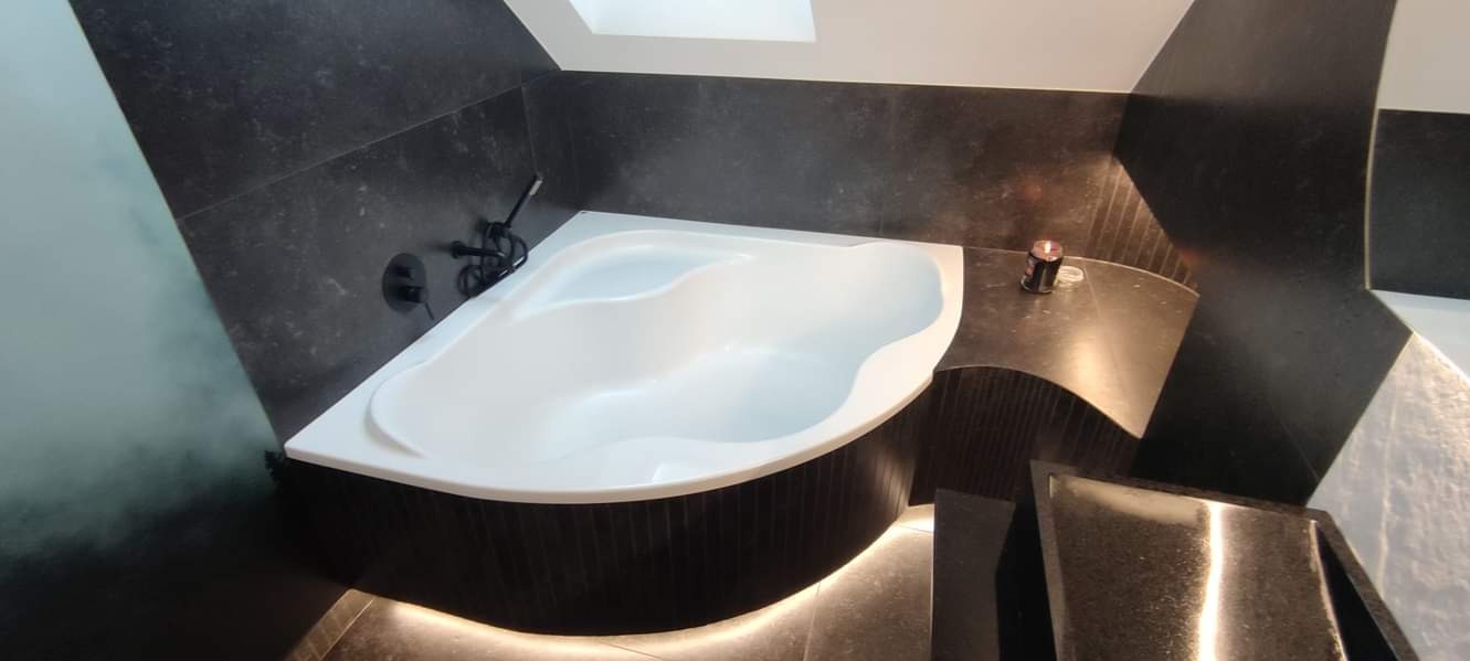 Narożna wanna z czarną armaturą na tle ciemnych płytek, oświetlona subtelnym światłem pod obudową, z zapaloną świeczką na blacie.