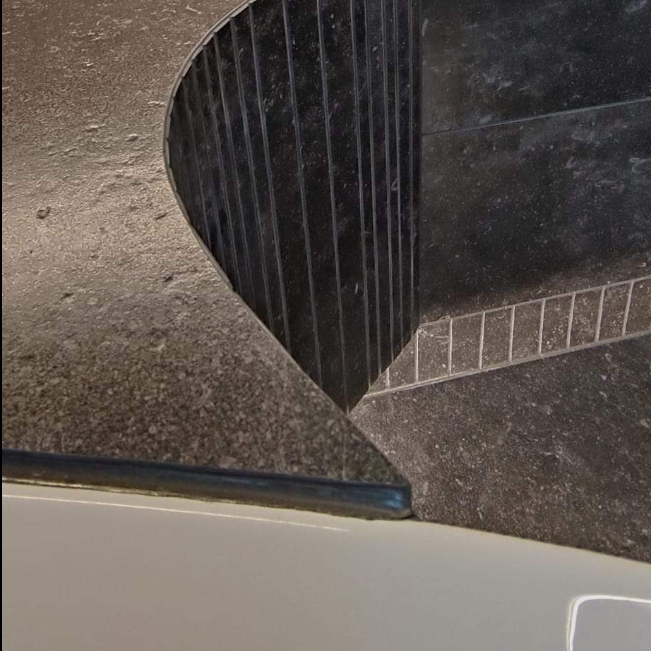 Fragment łazienki z ciemną, szorstką powierzchnią blatu i pionowymi, czarnymi listwami przy ścianie, widoczne białe wykończenie dolnej części.