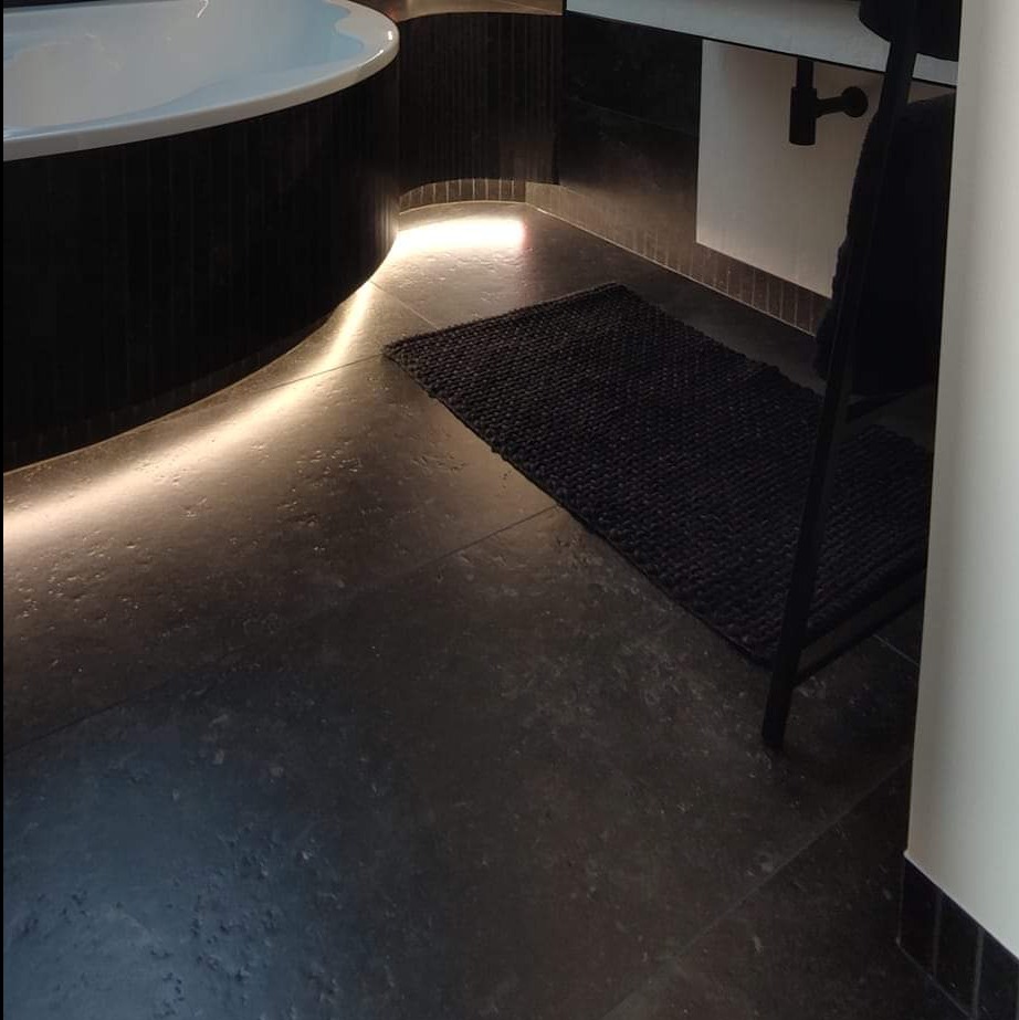 Ciemna, kamienna posadzka łazienki z podświetleniem LED przy wannie, czarny dywanik i fragment krzesła.