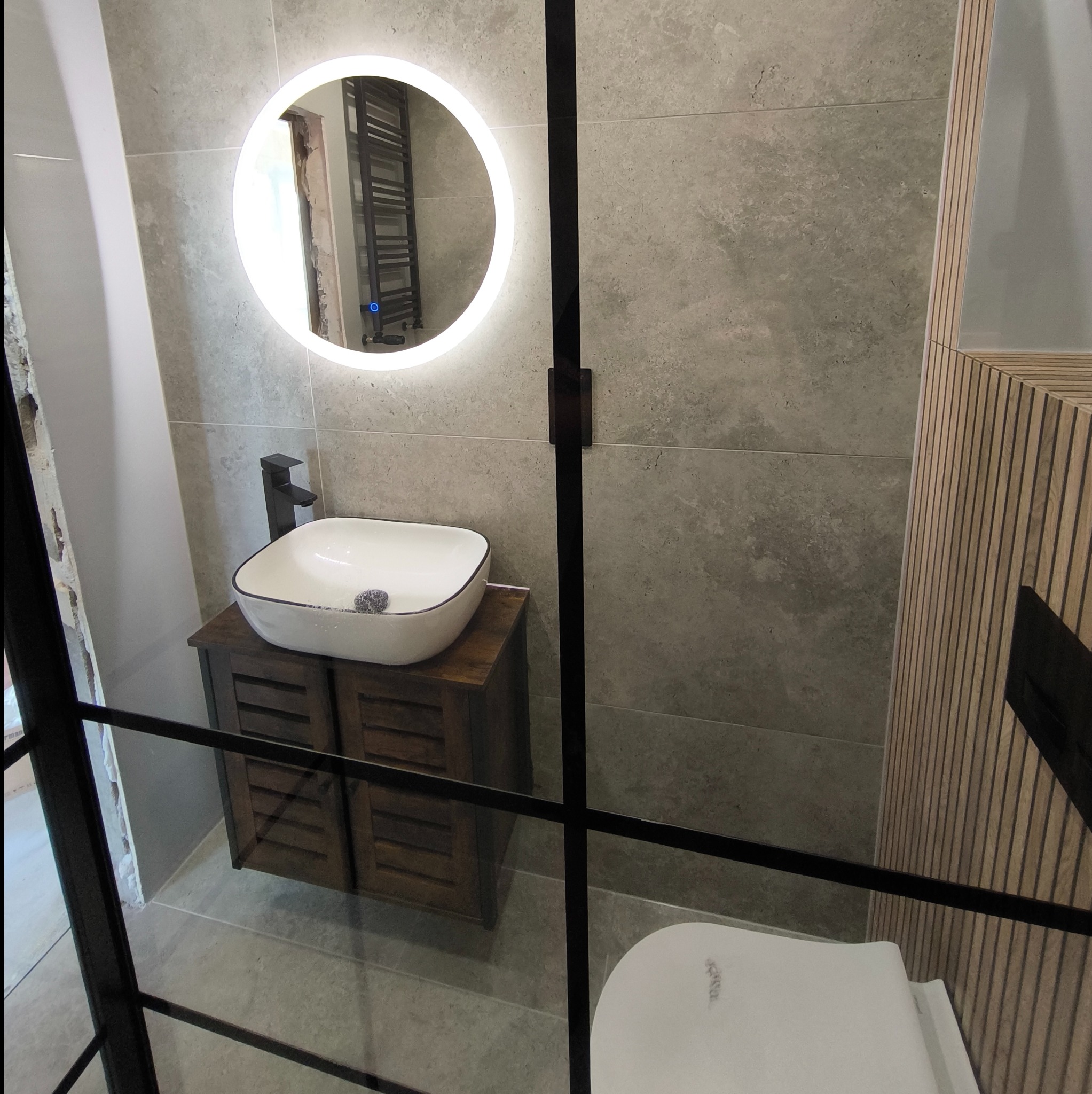 Nowoczesna łazienka z umywalką nablatową na szafce z ciemnego drewna, okrągłym lustrem z podświetleniem LED i szarą płytką imitującą kamień na ścianach, widok przez szklaną kabinę prysznicową...