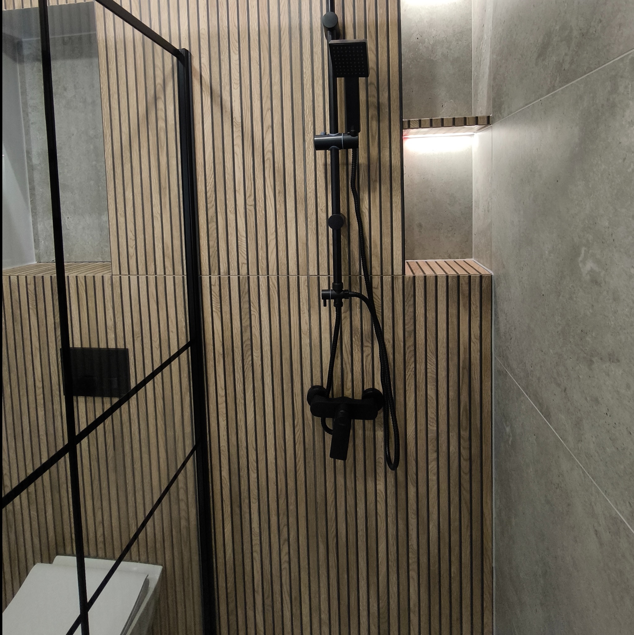 Nowoczesna łazienka z kabiną prysznicową typu walk-in, drewnianymi panelami ściennymi w pionowe paski i czarną armaturą, wnęka z podświetleniem LED.