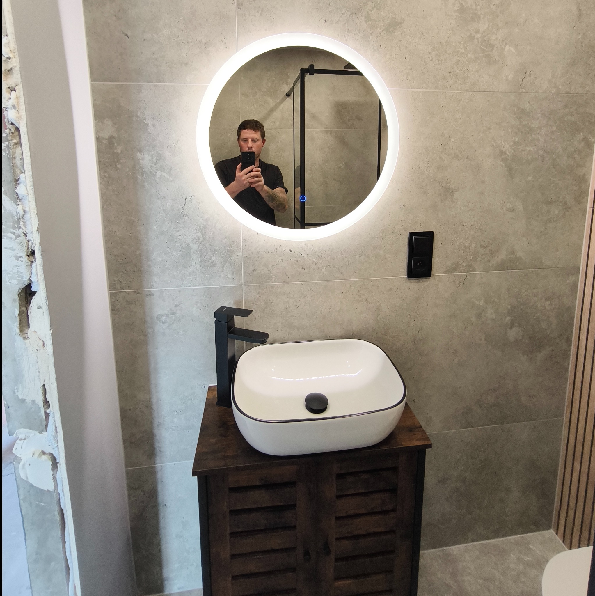 Umywalka nablatowa w łazience z okrągłym lustrem LED, widoczne szare płytki, czarna bateria i fragment ściany przed wykończeniem.