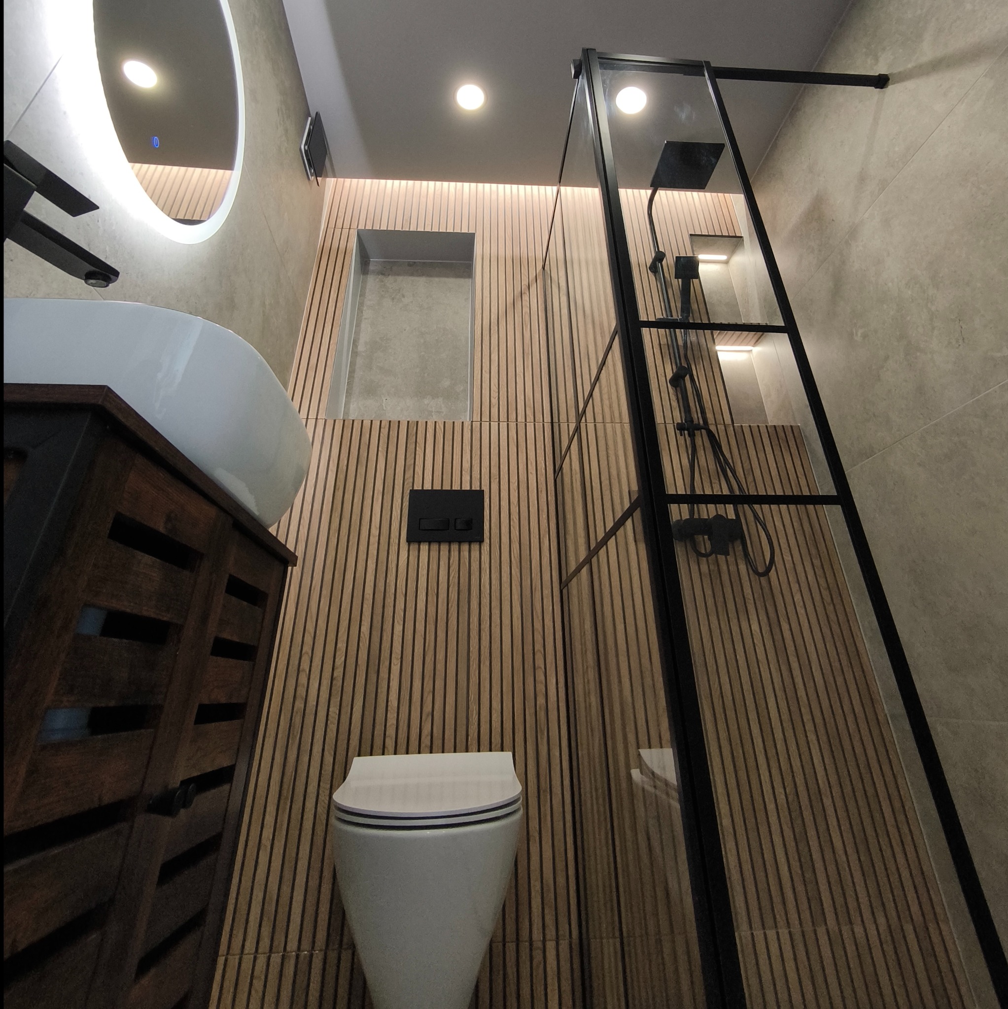 Nowoczesna łazienka z drewnianymi panelami ściennymi, okrągłym lustrem z podświetleniem LED, wiszącą miską WC i kabiną prysznicową z czarną ramą.