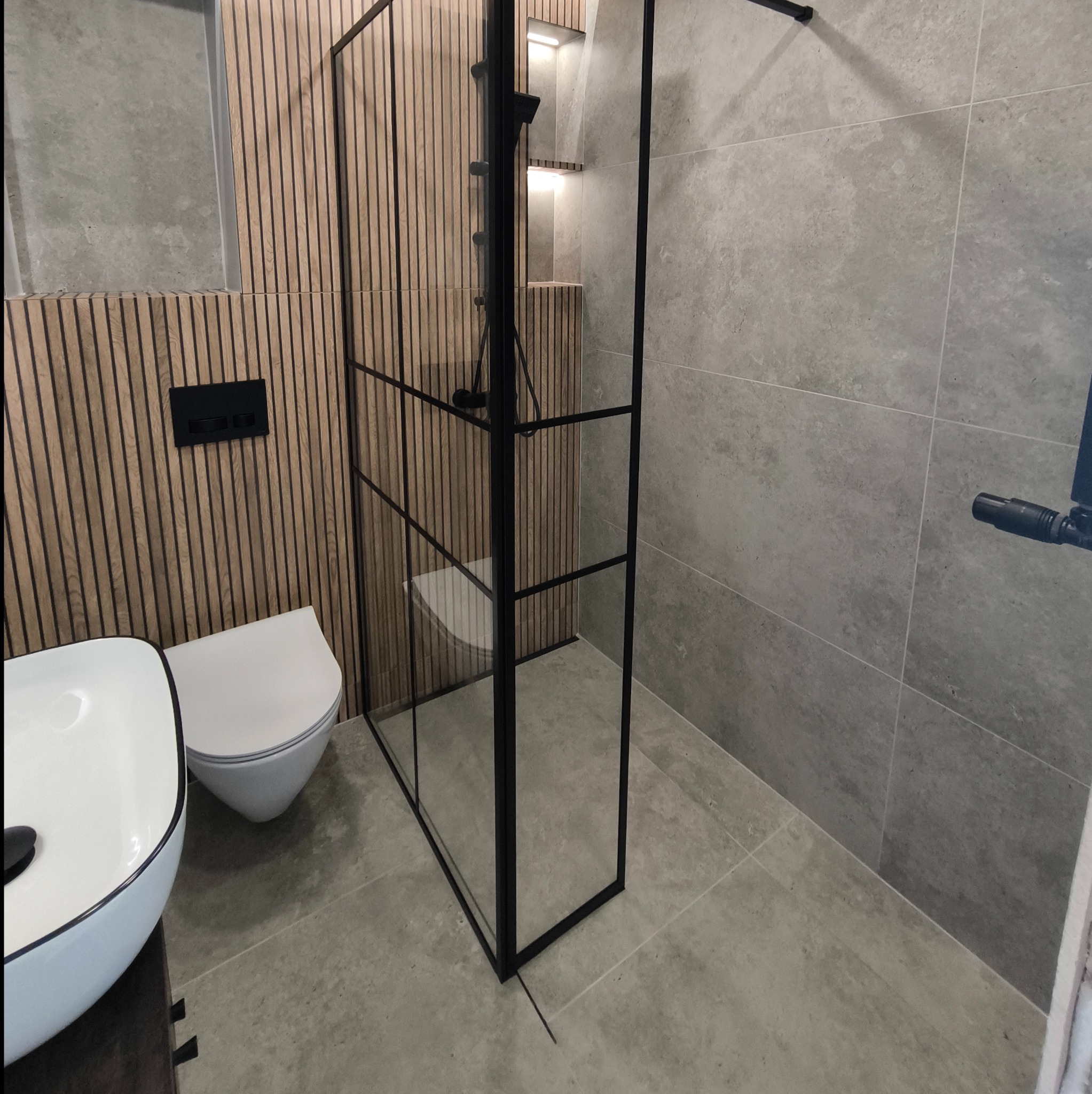 Nowoczesna łazienka z kabiną prysznicową typu walk-in z czarnymi profilami, umywalką nablatową i toaletą w zabudowie, wykończona płytkami imitującymi beton i drewnianymi lamelami na ścianie.