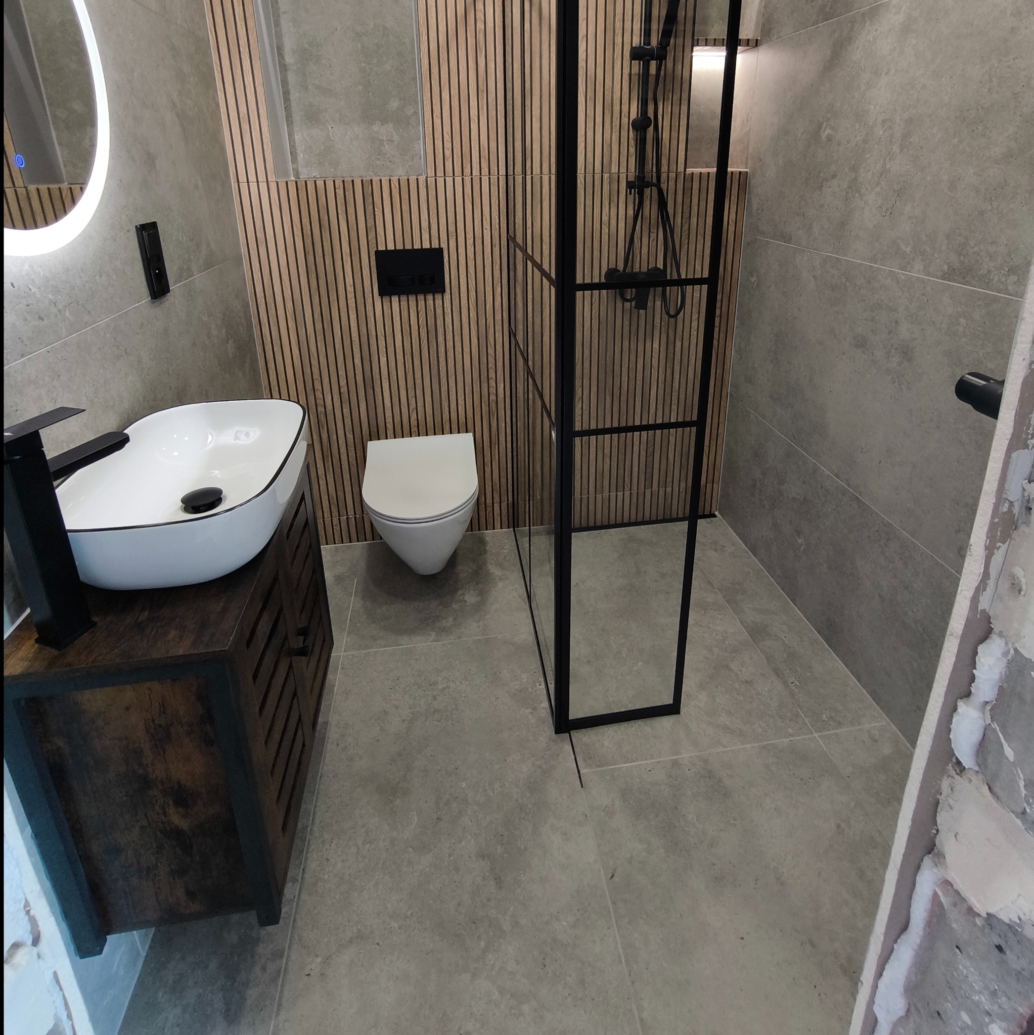 Nowoczesna łazienka z drewnianymi panelami ściennymi, umywalką nablatową na szafce z lamelami, toaletą podwieszaną i czarną kabiną prysznicową typu walk-in.