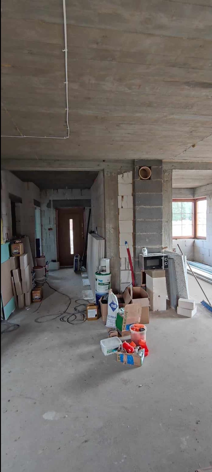 Wnętrze w stanie deweloperskim z widocznymi surowymi ścianami, betonowym stropem z poprowadzonymi kablami elektrycznymi, materiałami budowlanymi i narzędziami przygotowanymi do prac wykończeniowych.
