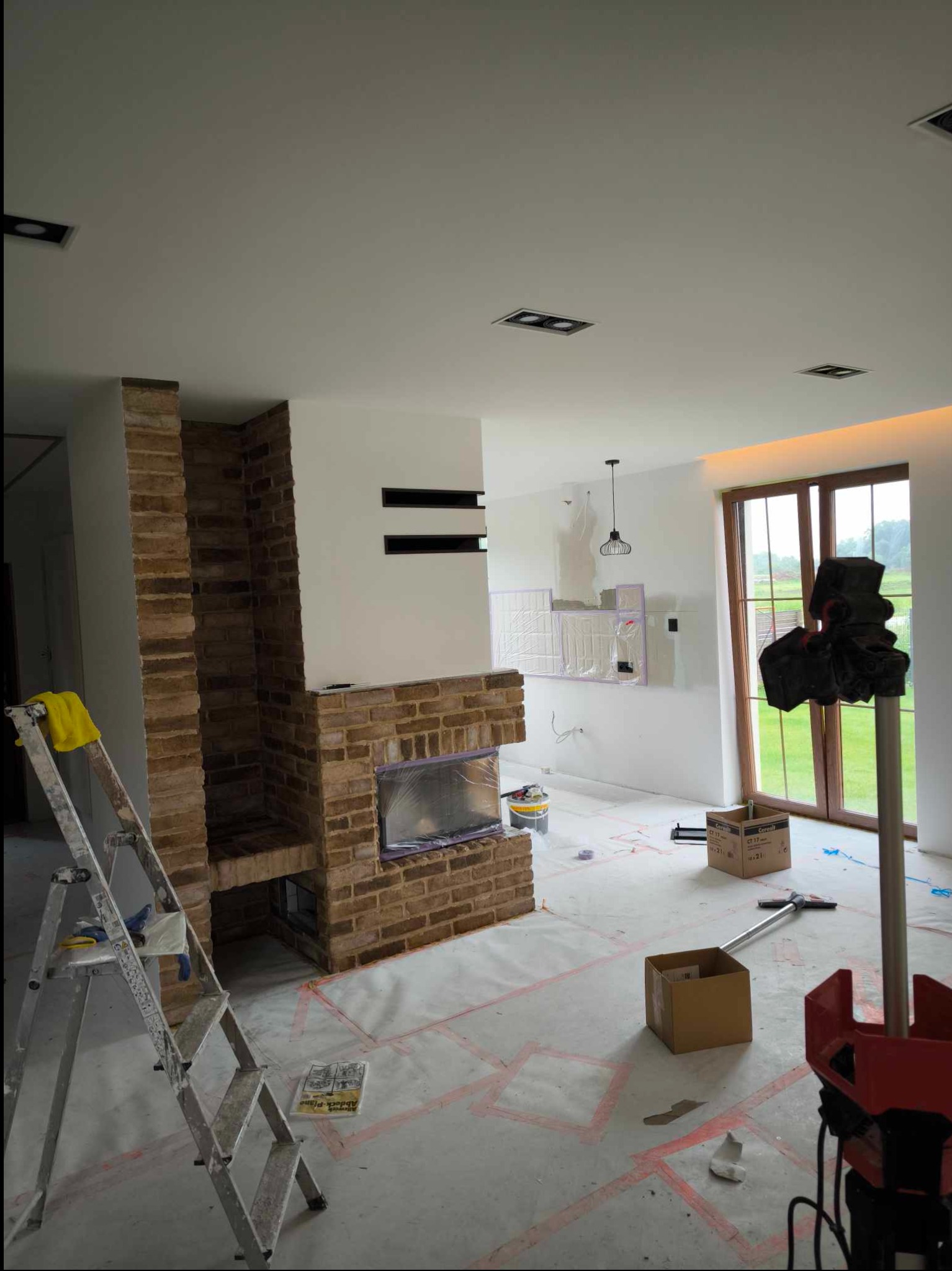 Salon w trakcie prac wykończeniowych: ceglany kominek z zabezpieczonym paleniskiem, białe ściany z czarnymi wnękami, zakryta folią podłoga, drabina aluminiowa z żółtą szmatką, narzędzia i kartony...