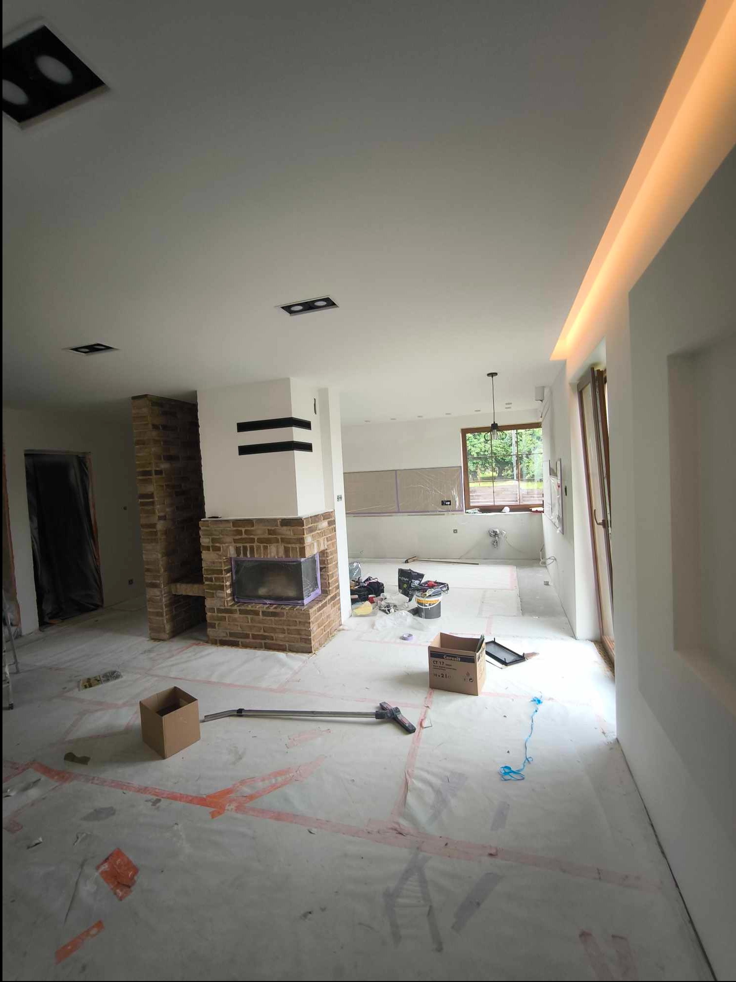 Przestronne wnętrze w trakcie prac wykończeniowych: zabezpieczona folią podłoga, ceglany kominek z czarnymi akcentami, puszki z farbą, narzędzia, oraz dekoracyjne oświetlenie sufitowe.