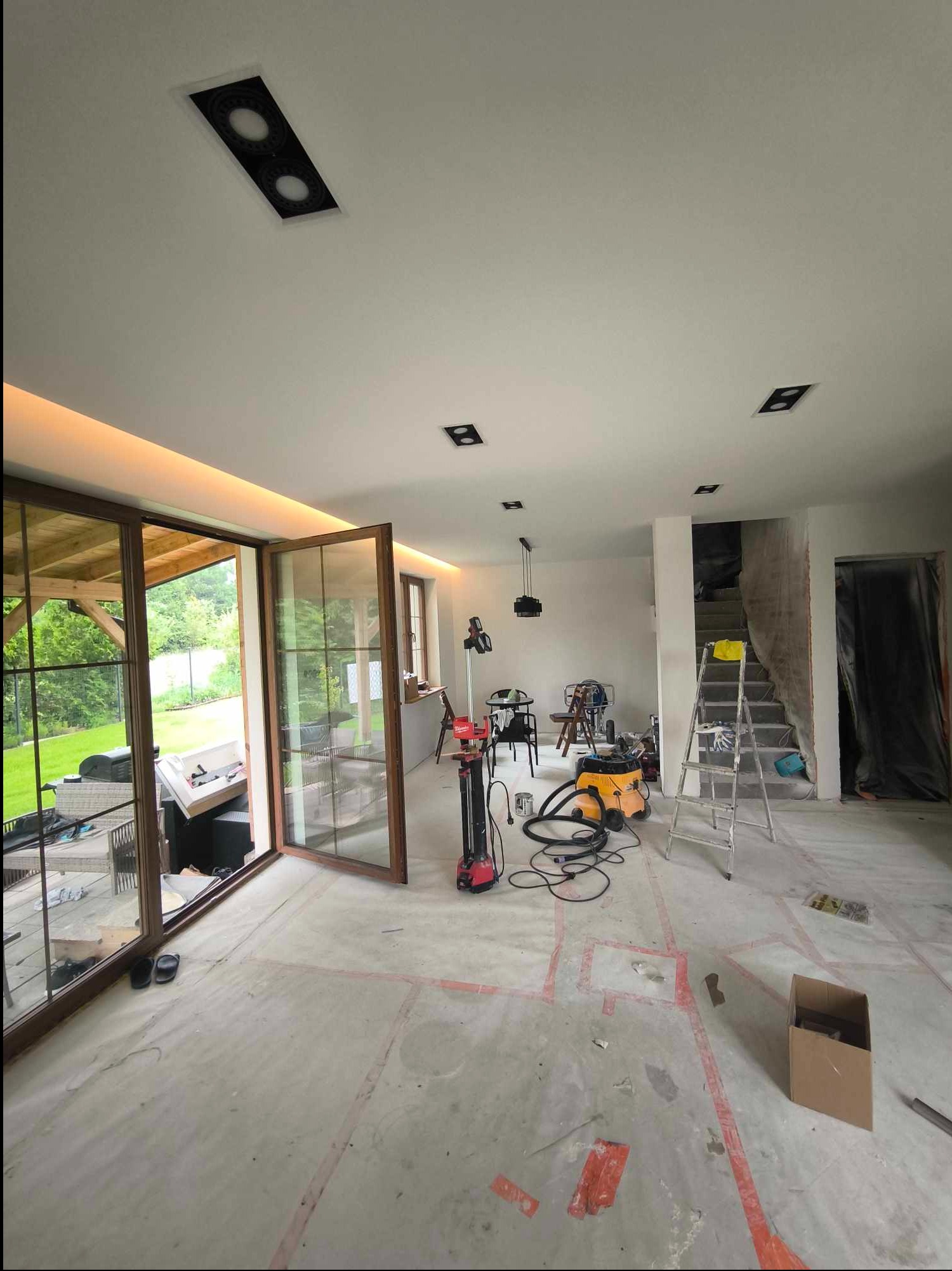 Wnętrze w trakcie prac wykończeniowych: otwarte przesuwne drzwi balkonowe z widokiem na zieleń, przygotowane podłoże, drabina, lampa wisząca, sprzęt budowlany i zakryte folią schody.