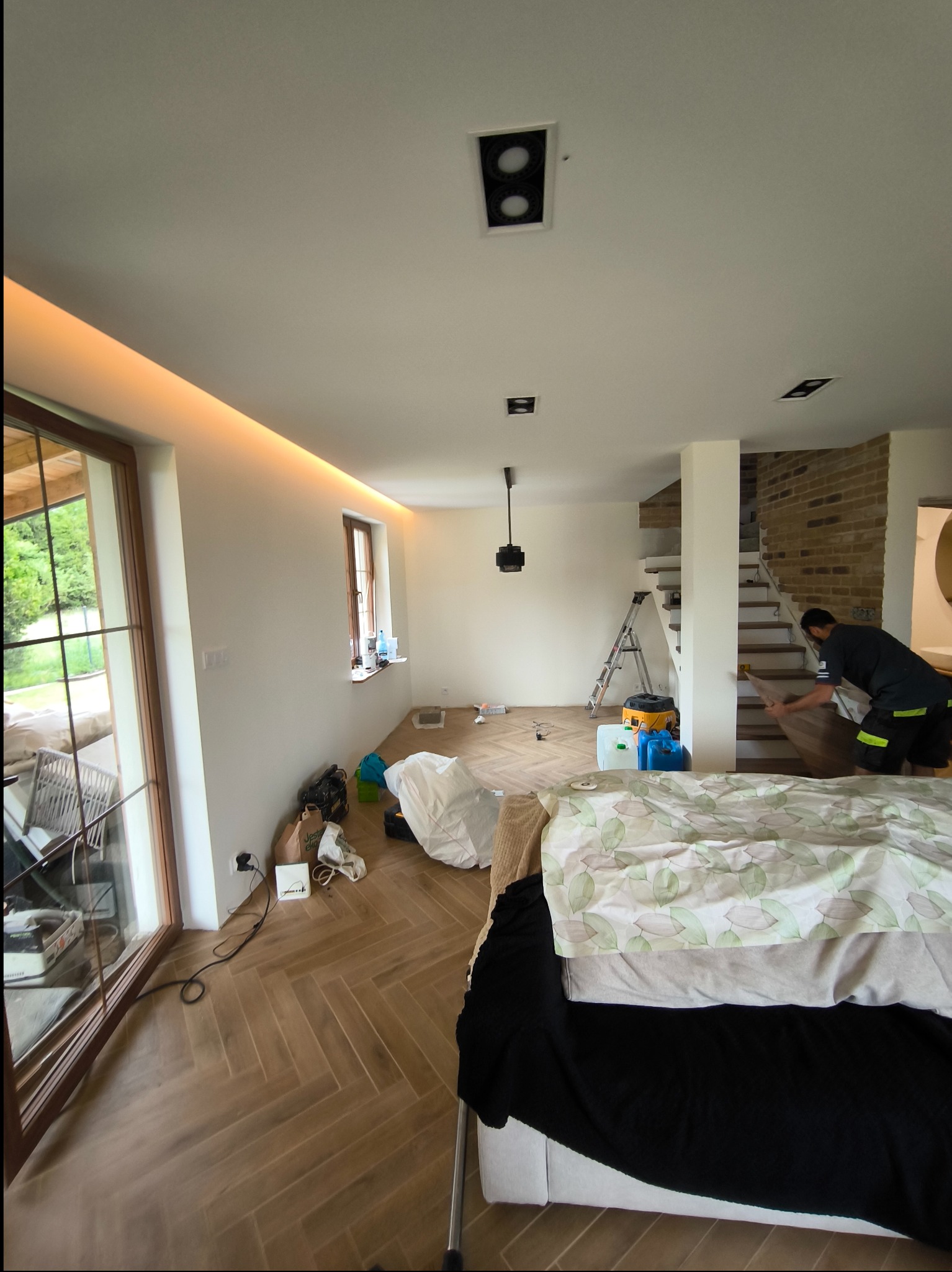 Montaż parkietu w jodełkę w jasnym wnętrzu z oknami i ceglaną ścianą, widoczny pracownik układający panele i drabina w tle.