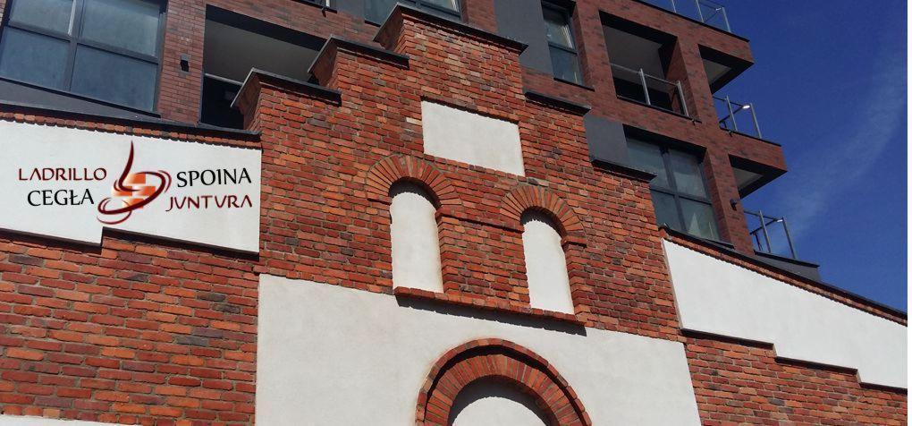 Fragment elewacji budynku z czerwonej cegły z dekoracyjnymi łukami i białymi elementami, widoczne logo firmy 'Ladrillo Cegła Spoina Jvntvra'.