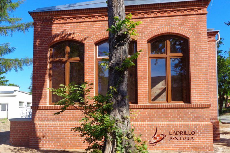 Elewacja budynku z czerwonej cegły z trzema łukowatymi oknami z brązowymi ramami, częściowo zasłonięta przez pień drzewa z zielonymi liśćmi. W tle widoczny fragment innego budynku.