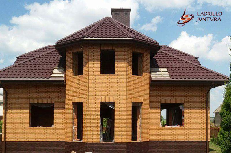 Niedokończony budynek z żółtej cegły z ciemnobrązowym dachem z blachodachówki, widoczne otwory okienne i drzwiowe, komin z cegły, logo firmy na górze po prawej.