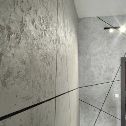 Finish Bud - Ściana z dekoracyjną, szarą strukturą, przedzielona czarnymi, cienkimi liniami, oświetlona listwą z czterema punktami świetlnymi, widoczny fragment drzwi.