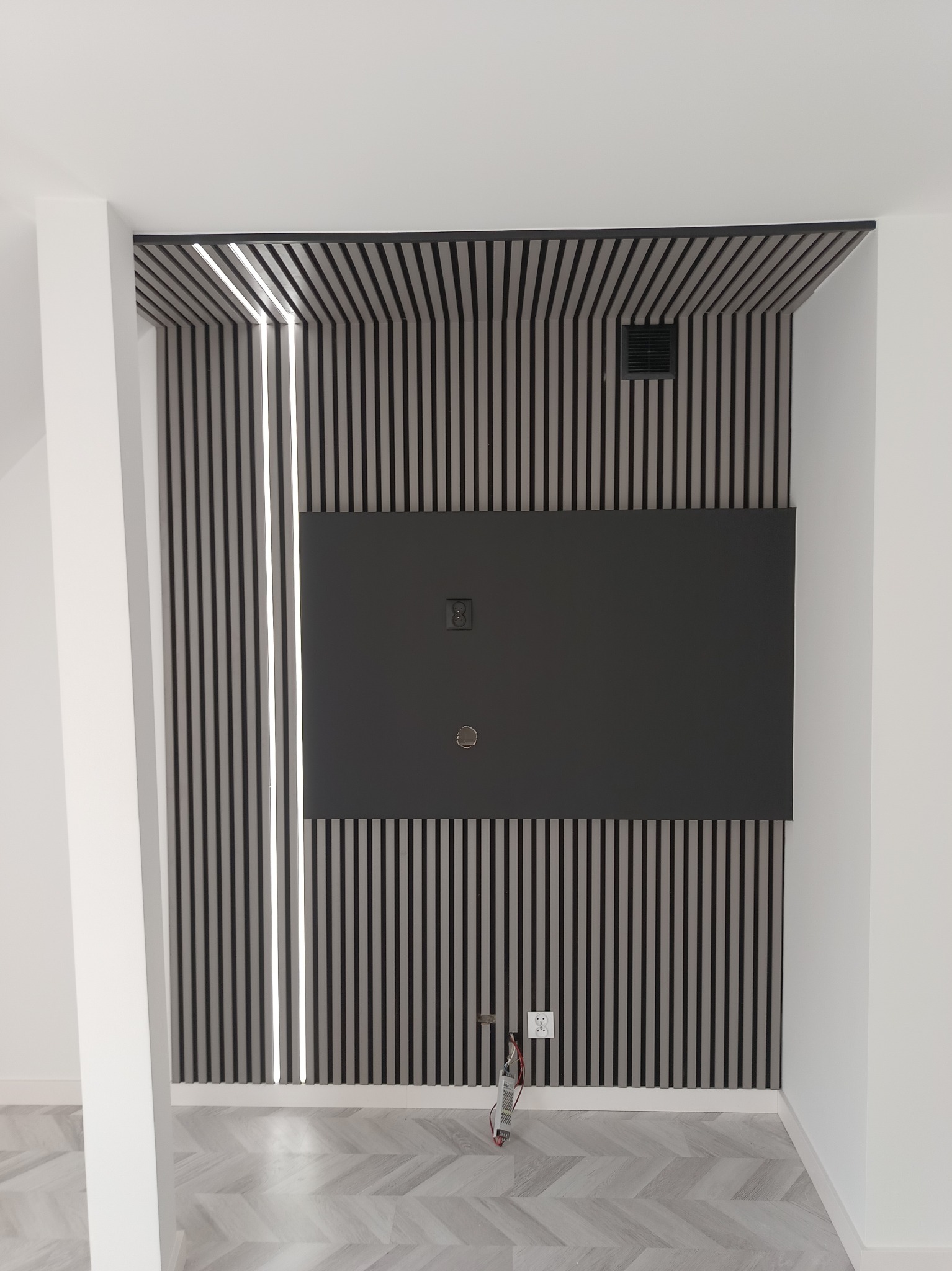 Nowoczesna ściana z paneli lamelowych w odcieniach szarości i czerni, podświetlona pionowymi paskami LED, z zamontowanym czarnym panelem centralnym z widocznymi otworami na kable i gniazdkiem...