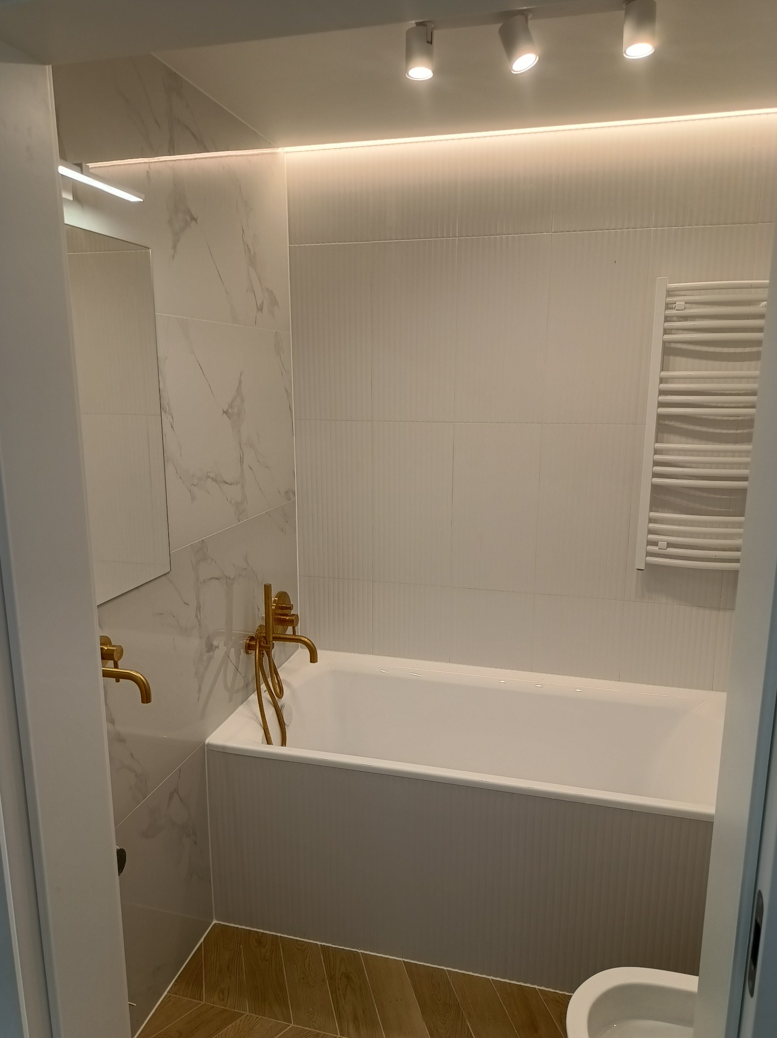 Nowoczesna łazienka z białą wanną, złotą armaturą i marmurowymi płytkami, oświetlona ledami i reflektorami.