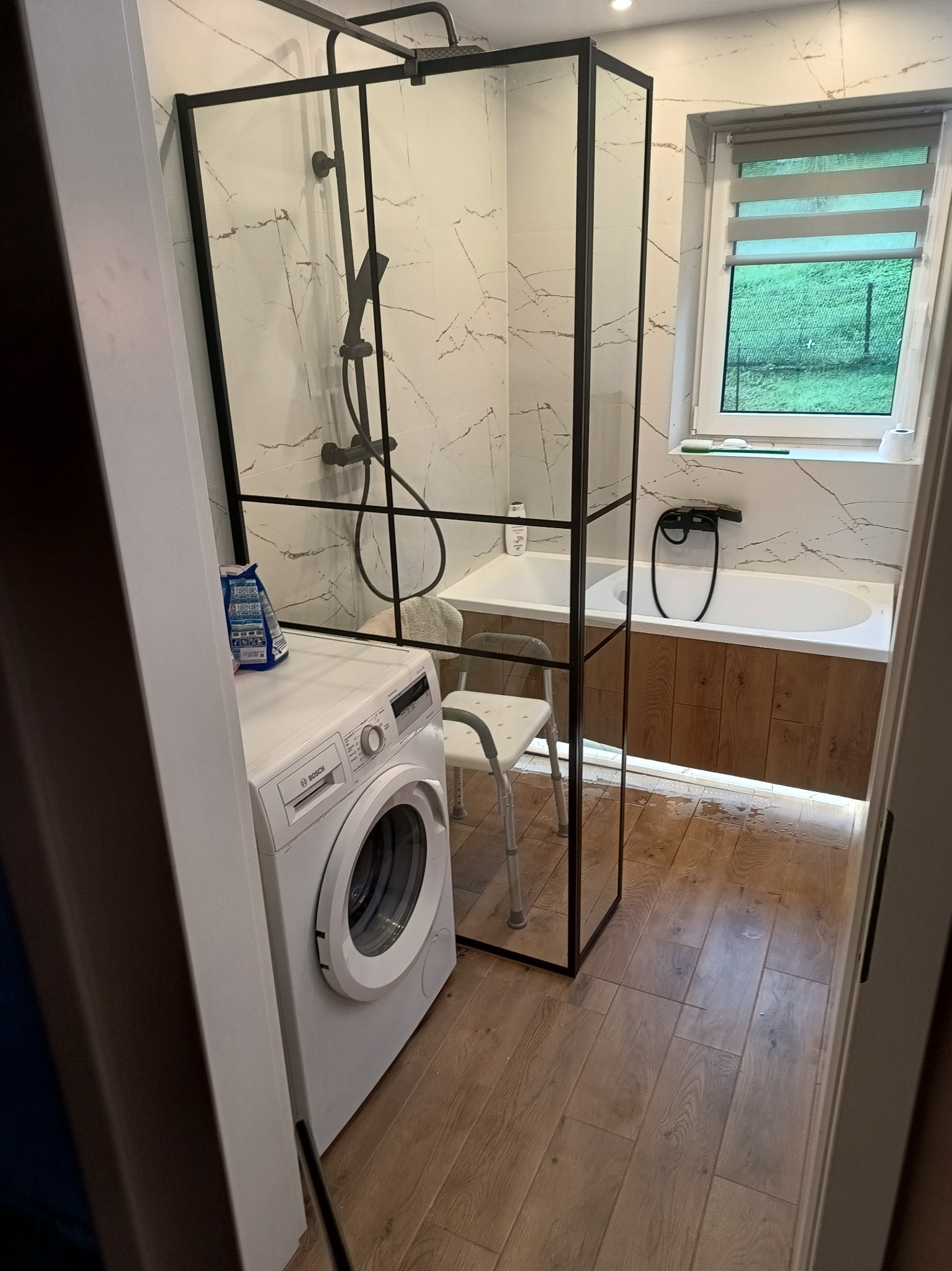 Nowoczesna łazienka z kabiną prysznicową typu walk-in z czarnymi profilami, białą pralką Bosch i drewnopodobną okładziną wanny, widok na okno z zielenią