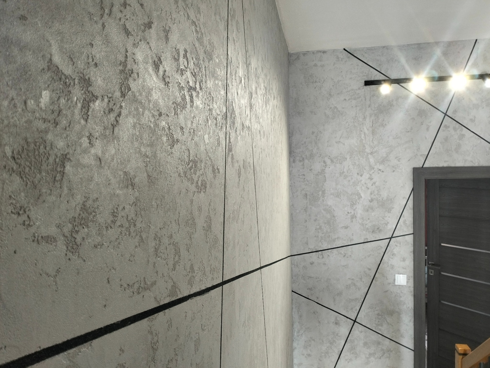 Ściana z dekoracyjną, szarą strukturą, przedzielona czarnymi, cienkimi liniami, oświetlona listwą z czterema punktami świetlnymi, widoczny fragment drzwi.