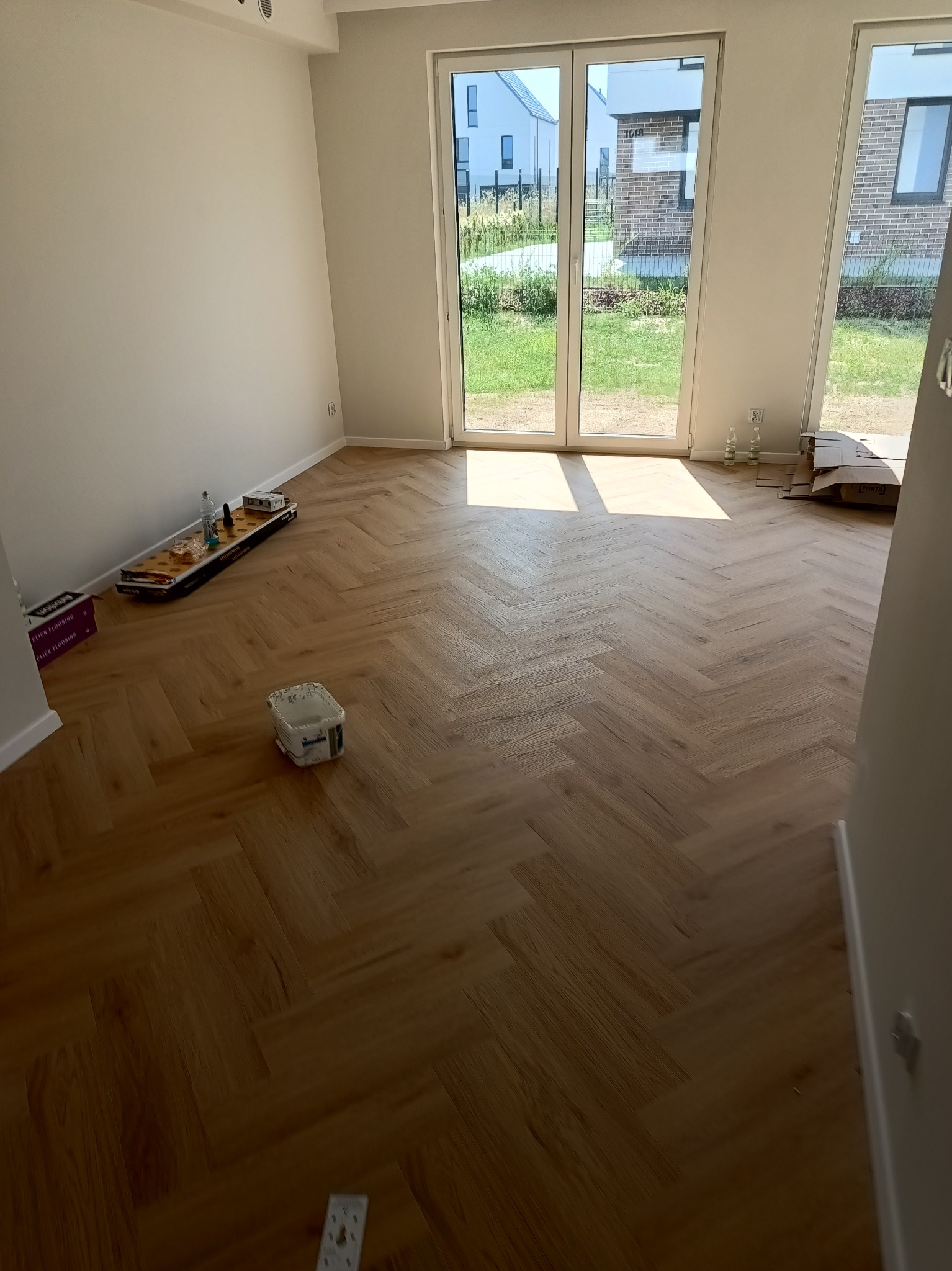 Ułożone panele podłogowe w jodełkę w jasnym, pustym pokoju z widokiem na ogród przez przeszklone drzwi balkonowe; widoczne puszki po farbie i materiały budowlane.