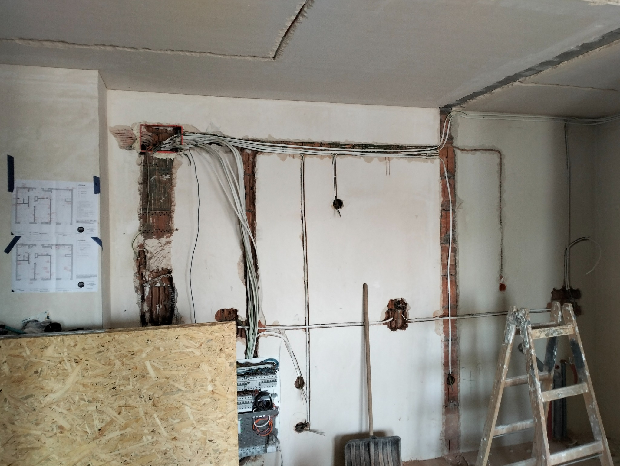 Wnętrze w trakcie prac instalacyjnych: odkryte przewody elektryczne w ścianach, skrzynka rozdzielcza, plan pomieszczenia na ścianie, drewniana płyta i drabina