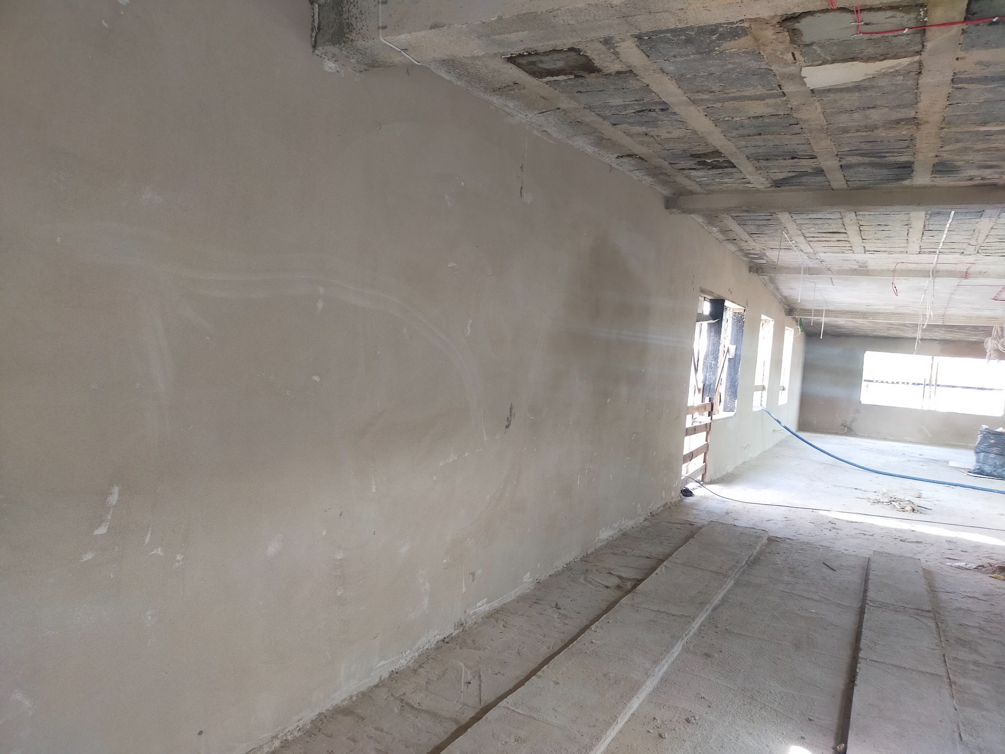 Ściana wewnętrzna po nałożeniu gładzi, widoczne okna i surowy betonowy sufit z wystającymi przewodami elektrycznymi.