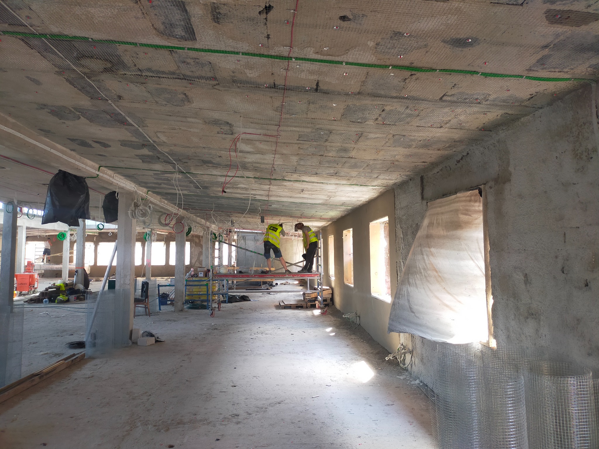 Wnętrze w trakcie prac wykończeniowych: dwóch robotników na rusztowaniu, montaż instalacji pod sufitem, widoczne przewody, siatka zabezpieczająca, okna zabezpieczone folią.