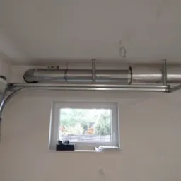 Widok na system kanałów wentylacyjnych pod sufitem w garażu, część kanałów zaizolowana folią aluminiową, widoczne okno z widokiem na zewnątrz.