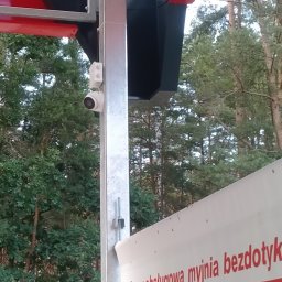 INSTALACJE Radosław Komosa - Kamera monitoringu na konstrukcji myjni bezdotykowej, widok z dołu. Tło stanowią drzewa. Szara metalowa konstrukcja, biała kamera.