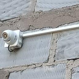 INSTALACJE Radosław Komosa - Kamera monitoringu IP zamontowana na szarej, surowej ścianie z cegieł. Widoczny kabel i biała listwa montażowa. Ujęcie z bliska, lekko z dołu.