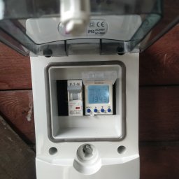 INSTALACJE Radosław Komosa - Biała skrzynka z otwartą pokrywą, wewnątrz wyłącznik Eaton i programator czasowy z wyświetlaczem LCD, zamontowana na drewnianej ścianie. Widoczne oznaczenia IP65 i CE.