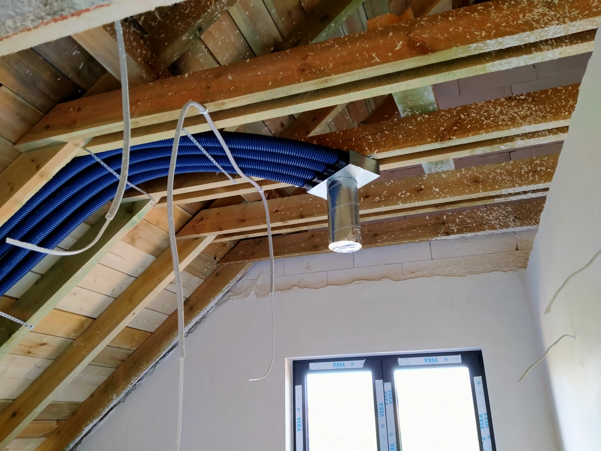Niebieskie rury wentylacyjne przymocowane do drewnianej konstrukcji dachu na poddaszu, widoczny metalowy element systemu wentylacyjnego i okno dachowe.