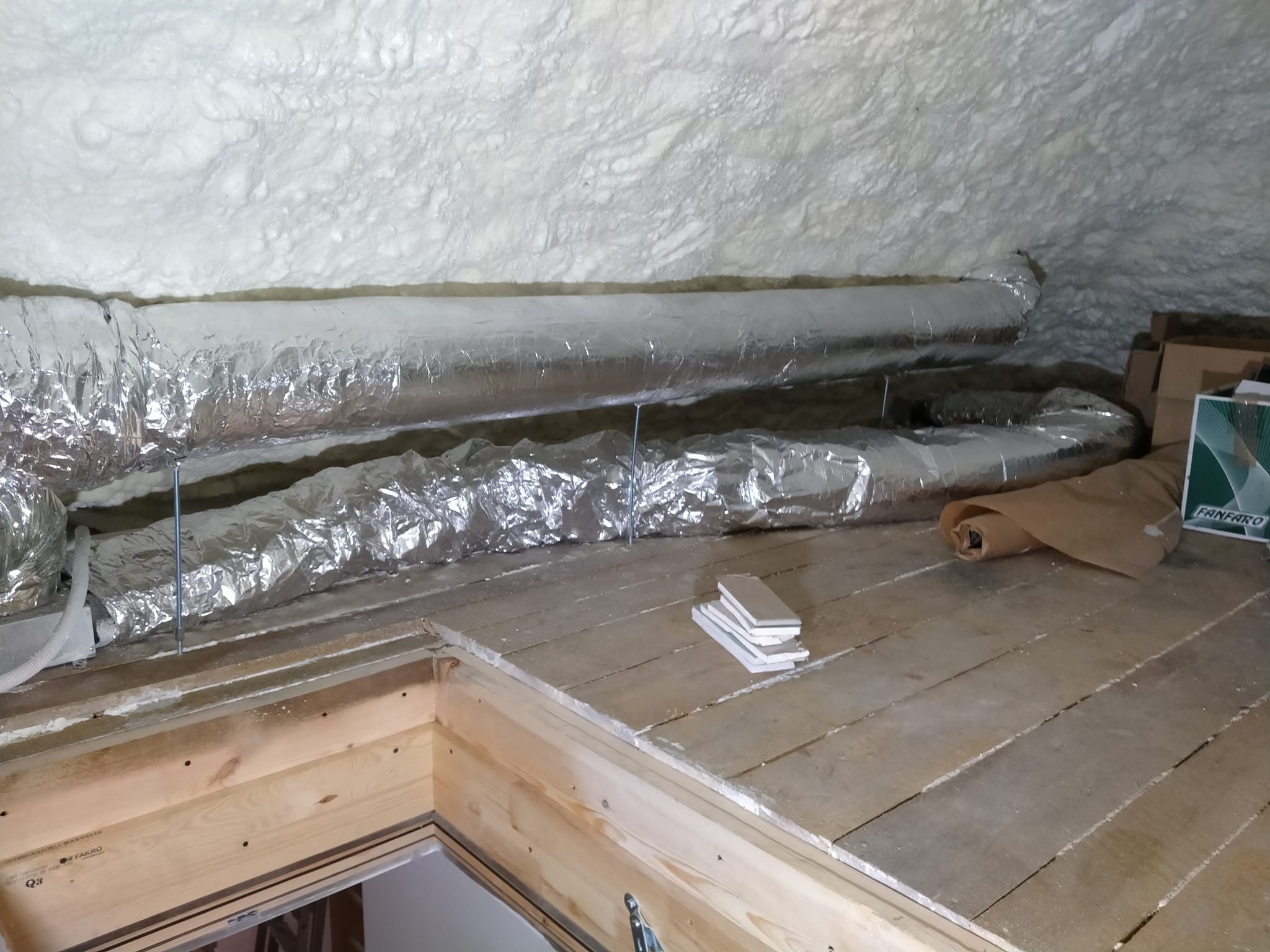 Izolacja poddasza pianką PUR z widocznymi przewodami wentylacyjnymi w srebrnej folii i fragmentem drewnianej podłogi z materiałami budowlanymi.