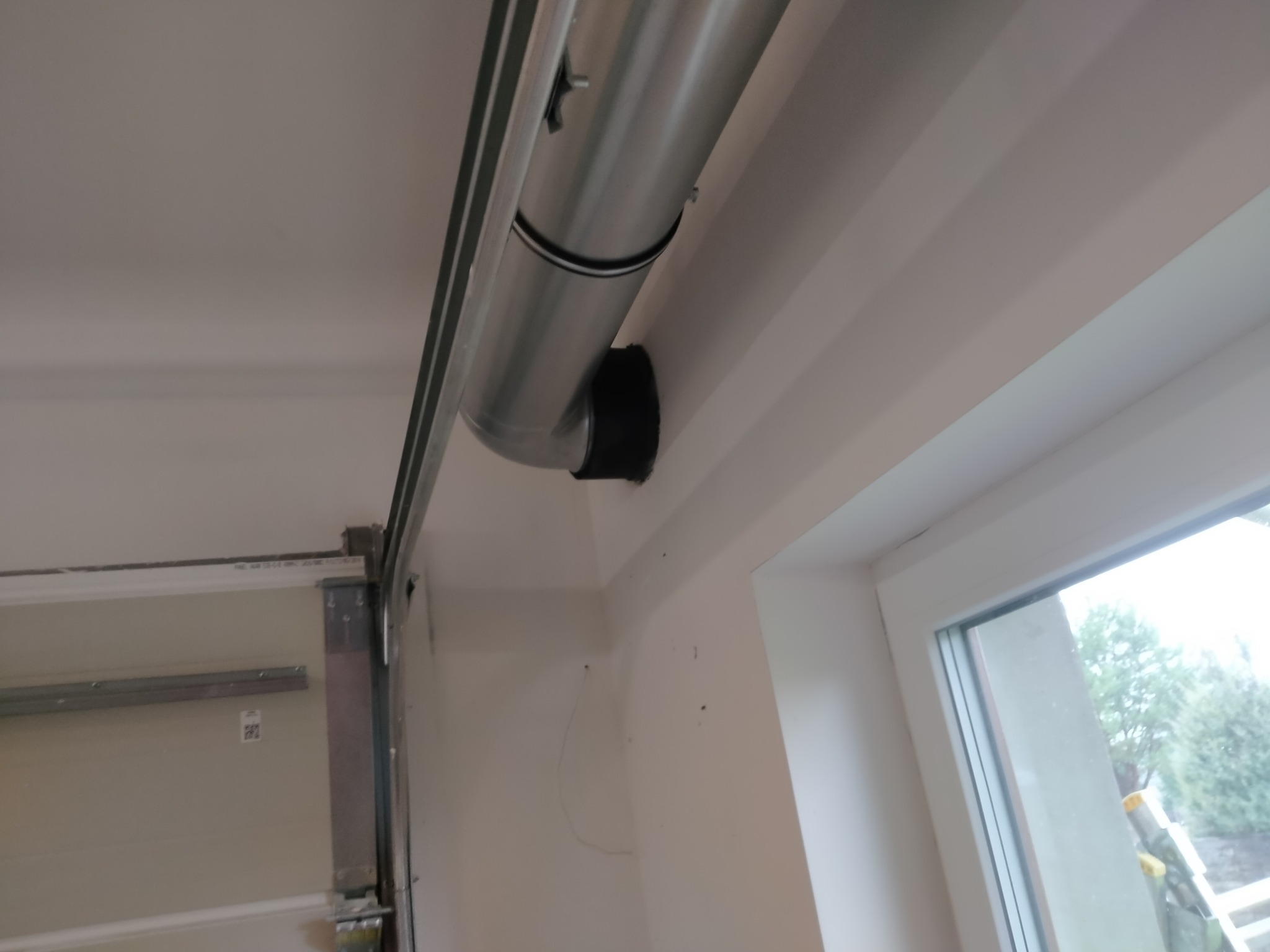 Szary kanał wentylacyjny z czarną uszczelką przymocowany do sufitu w pobliżu okna i ramy bramy garażowej.