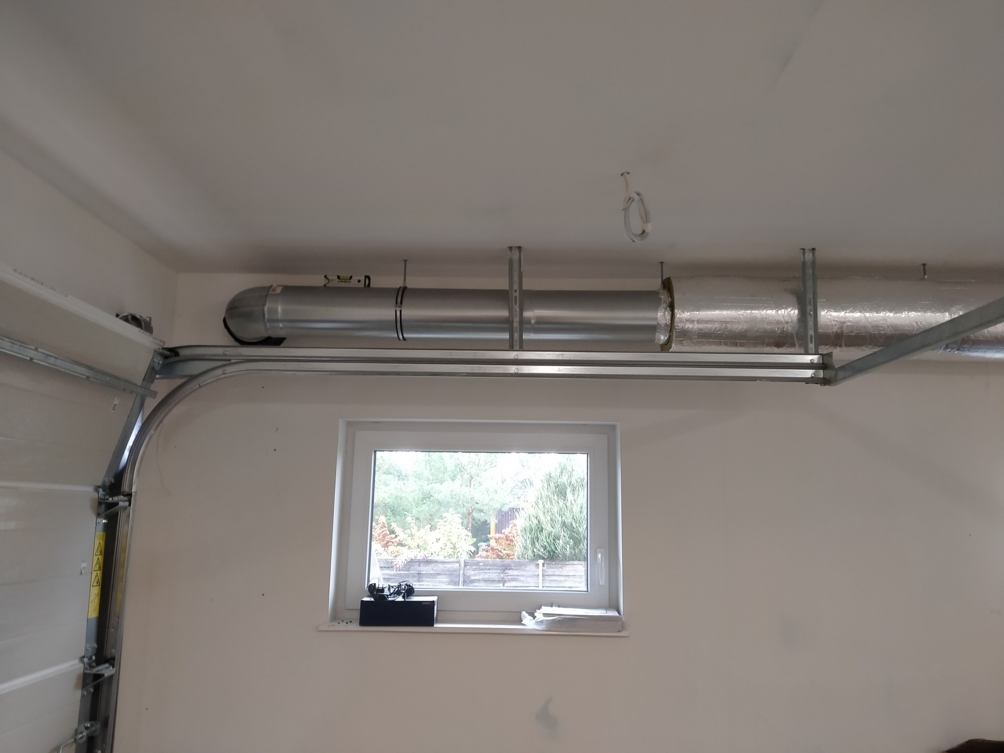 Widok na system kanałów wentylacyjnych pod sufitem w garażu, część kanałów zaizolowana folią aluminiową, widoczne okno z widokiem na zewnątrz.