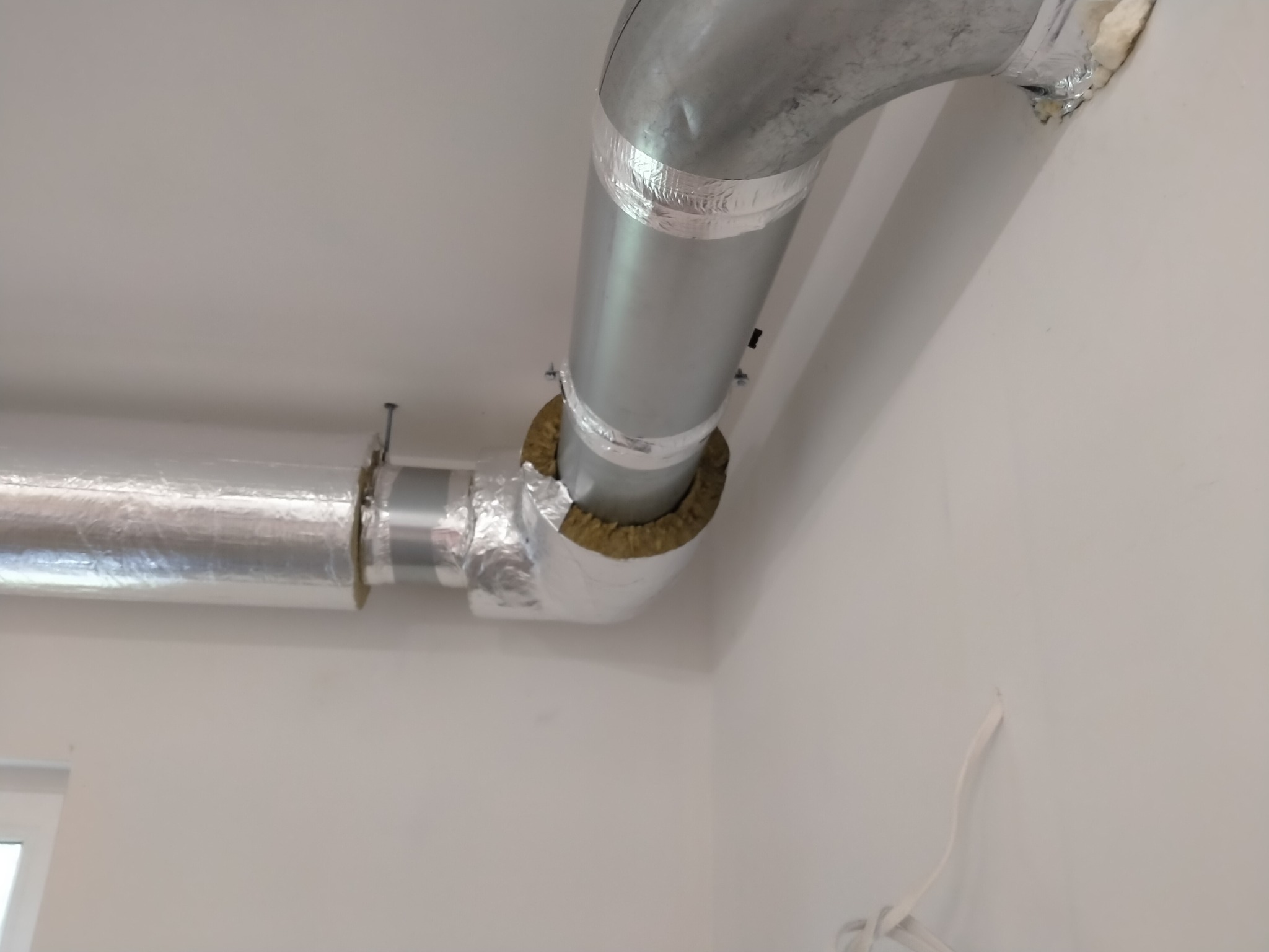 Metalowe rury wentylacyjne pod sufitem, zaizolowane wełną mineralną i owinięte folią aluminiową, widoczne mocowania do sufitu.