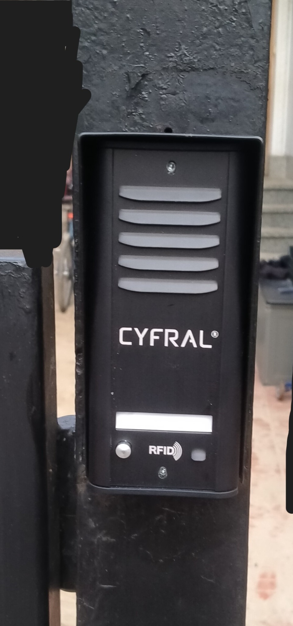 Czarny wideodomofon Cyfral z czytnikiem RFID zamontowany na metalowym słupku bramy. Widoczne ślady użytkowania na obudowie i słupku.
