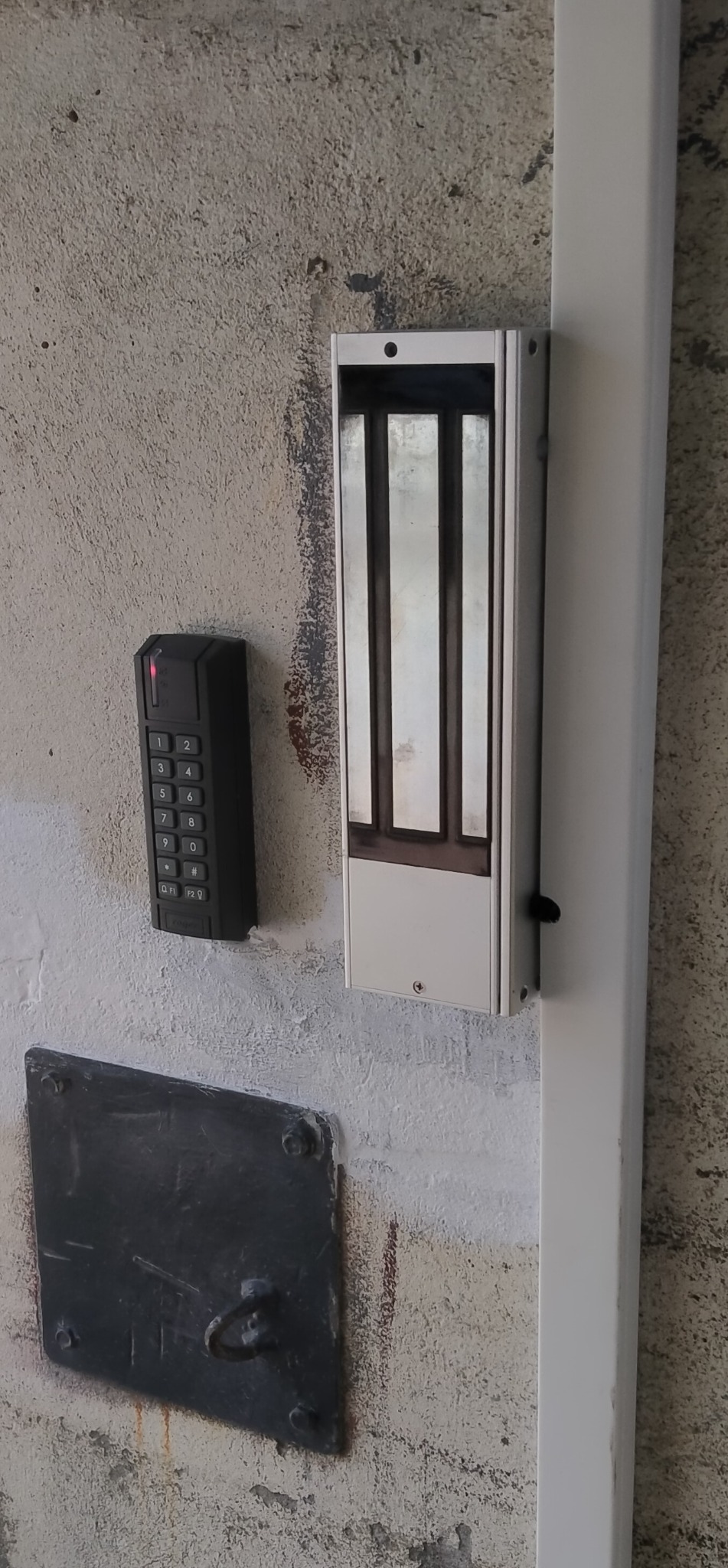Zamek elektromagnetyczny z klawiaturą kodową na szorstkiej ścianie. Widoczna metalowa listwa i stara, metalowa płytka z uchwytem. Zabezpieczenie dostępu.