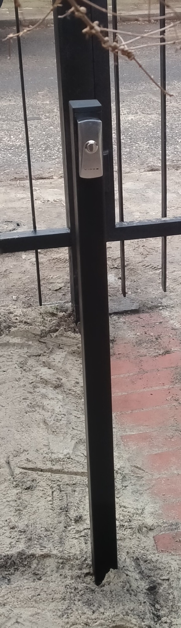 Zamontowany wideodomofon Vidos na czarnej bramie posesji w Ostrołęce. Widoczny fragment muru i gałęzie drzewa. Ujęcie z bliska, skupione na urządzeniu.