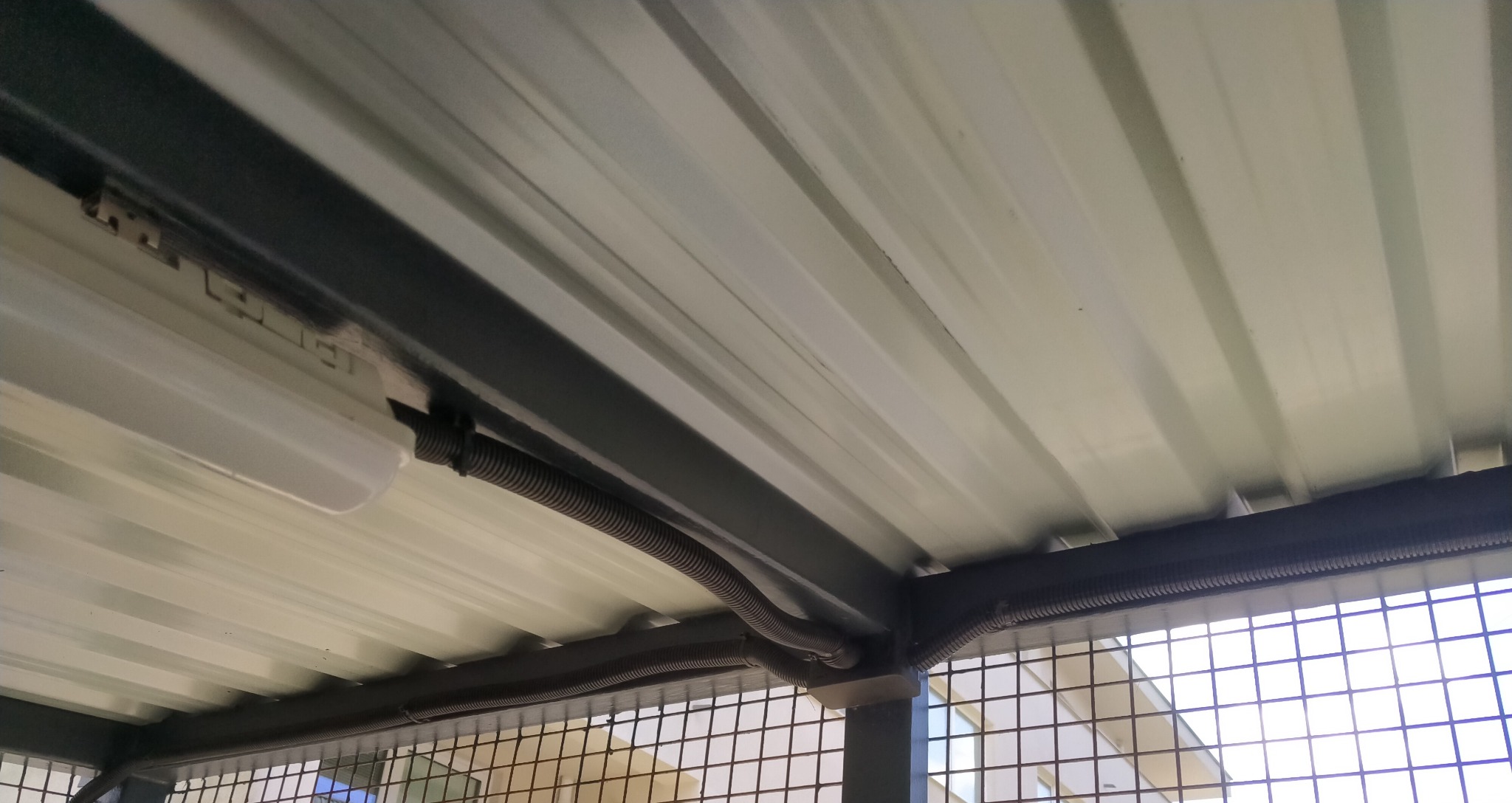 Instalacja elektryczna w peszlu pod blachą trapezową, widoczna lampa LED i metalowa konstrukcja. Ujęcie z dołu, widok na sufit.