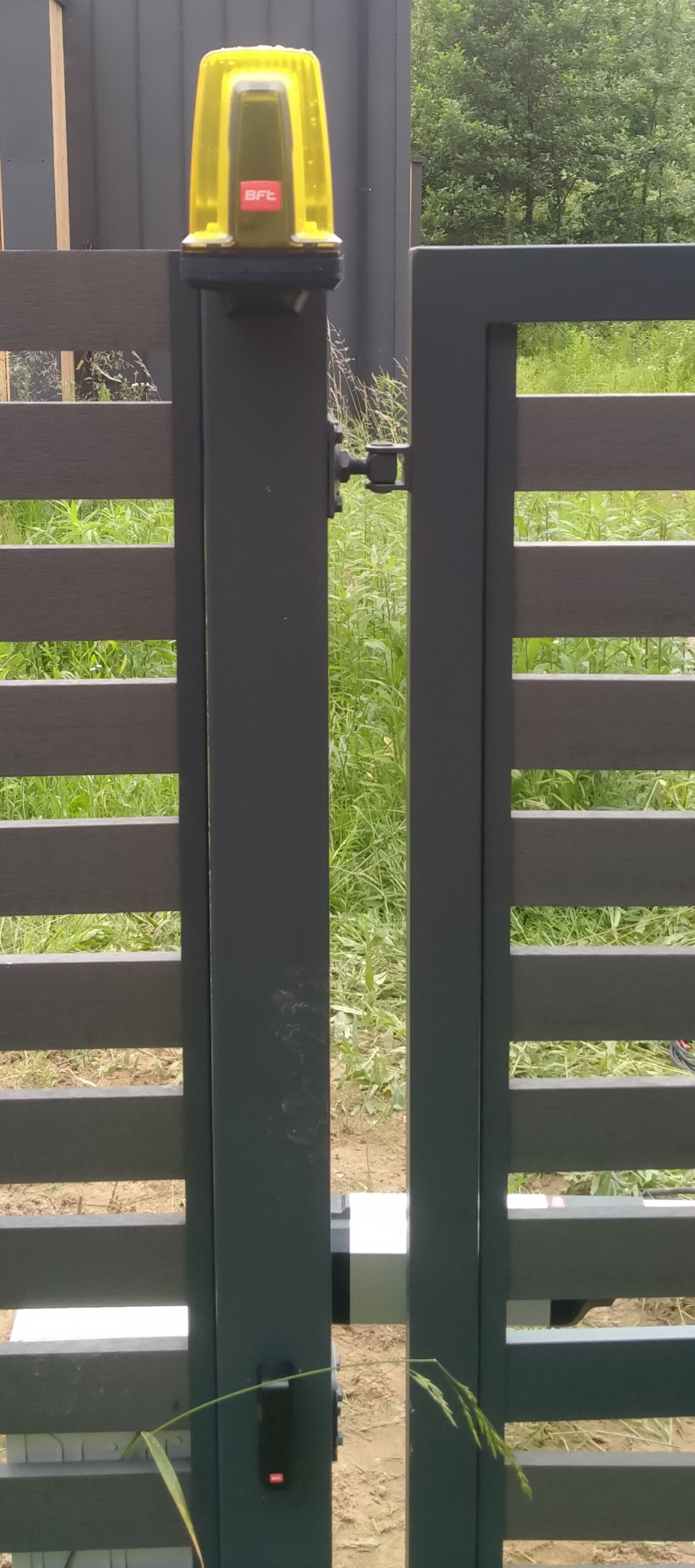 Nowoczesna brama z automatyką BFT. Widoczny siłownik, lampa ostrzegawcza i czytnik kart. Tło: zieleń ogrodu. Ujęcie z bliska, prezentujące detale instalacji.