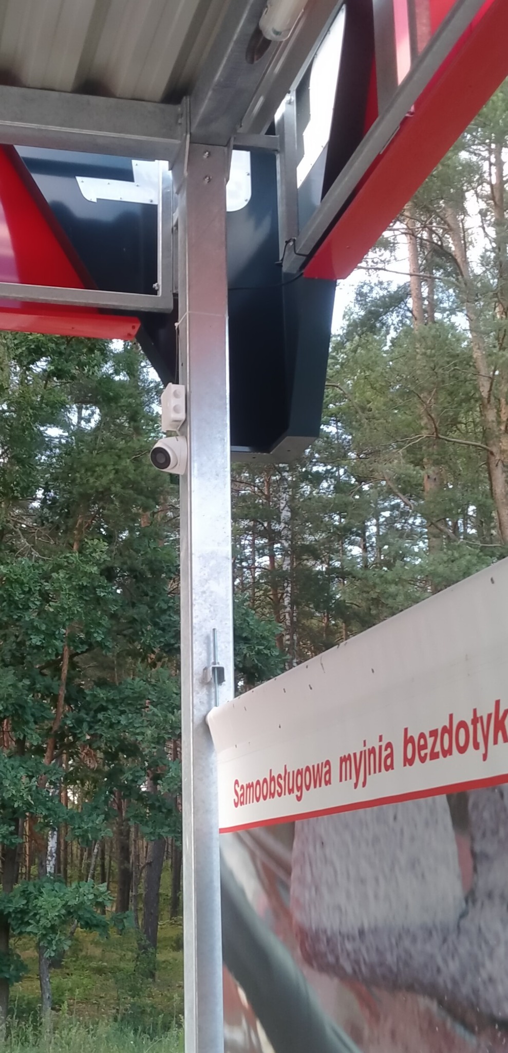Kamera monitoringu na konstrukcji myjni bezdotykowej, widok z dołu. Tło stanowią drzewa. Szara metalowa konstrukcja, biała kamera.