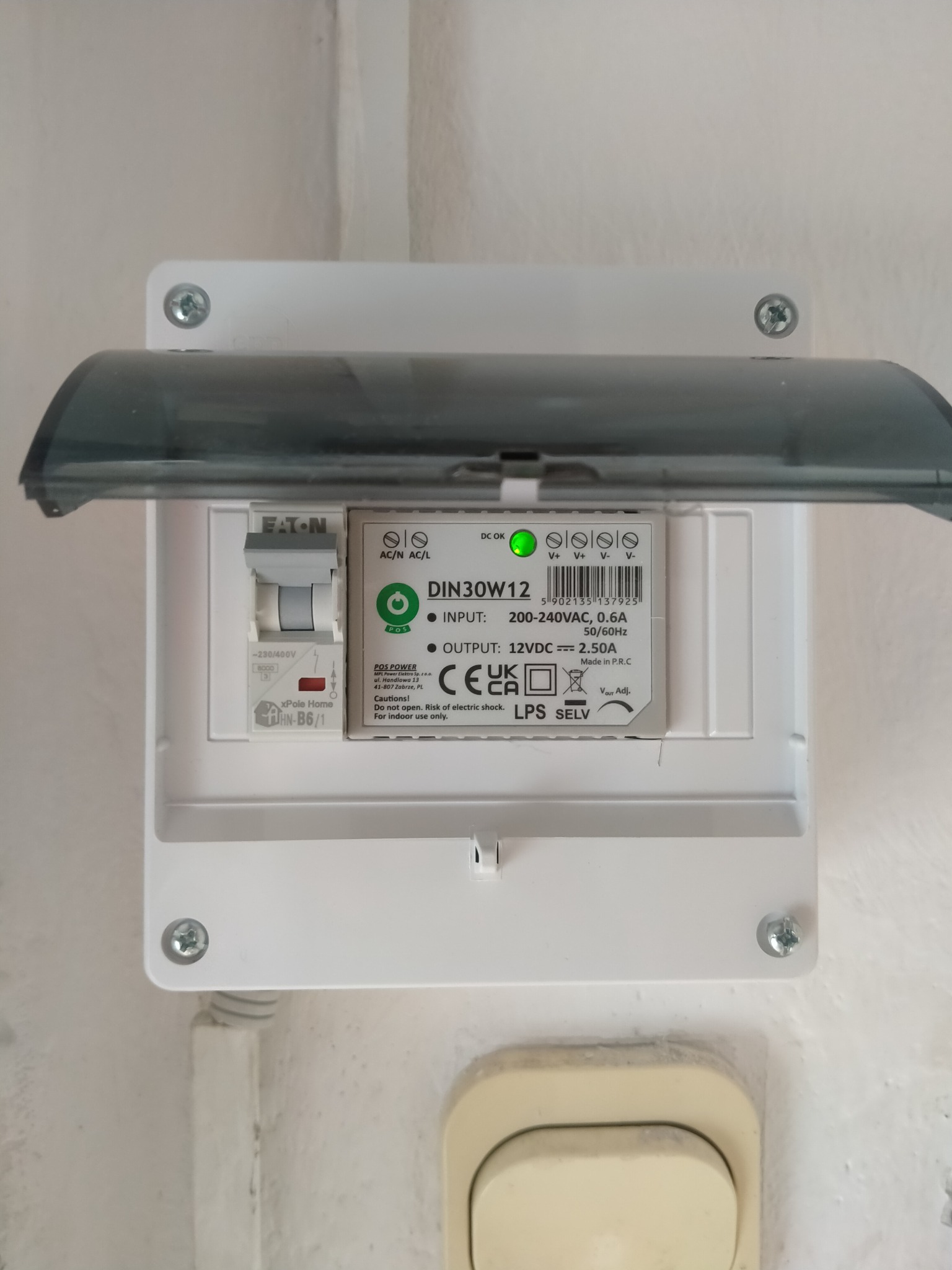 Otwarta skrzynka zasilająca Eaton DIN30W12 z zieloną diodą LED, bezpiecznikiem xPole Home i informacjami o napięciu. Widoczne śruby mocujące i fragment ściany.
