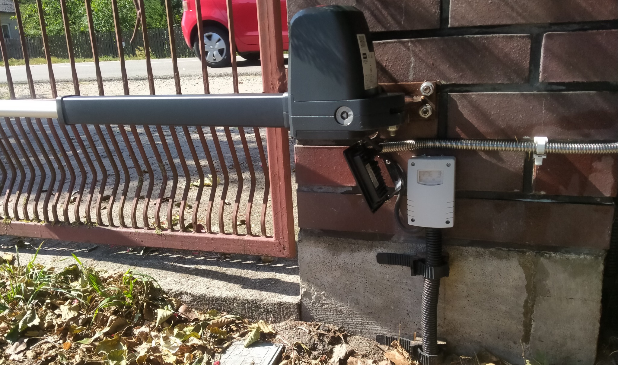 Zamontowany napęd bramy skrzydłowej z czujnikiem ruchu na ceglanym słupku. Widoczny fragment bramy i czerwony samochód w tle. Oświetlenie dzienne.