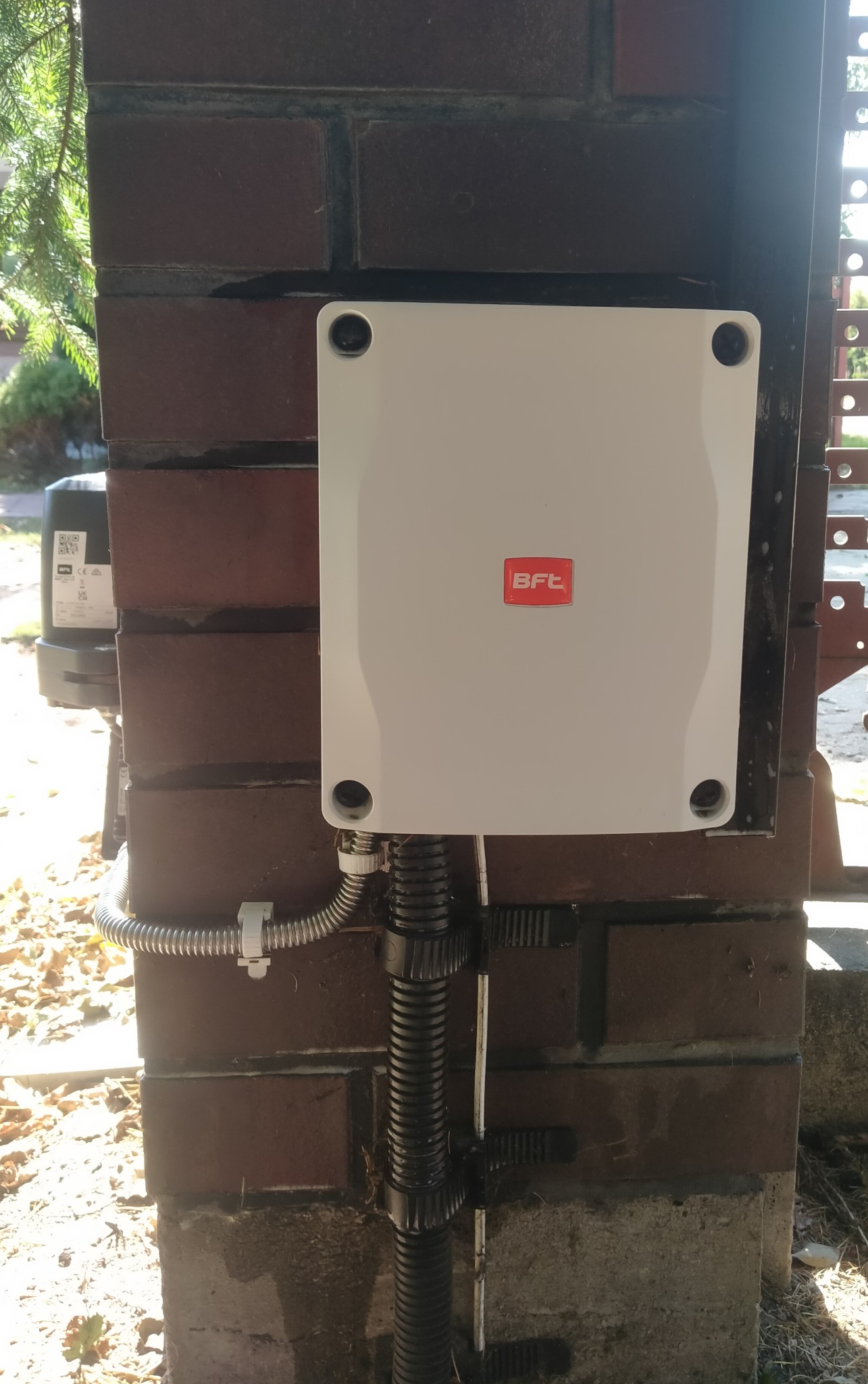 Biała skrzynka sterownicza BFT zamontowana na ceglanym słupku bramy, z widocznymi przewodami w peszlu. Widoczny fragment napędu bramy.