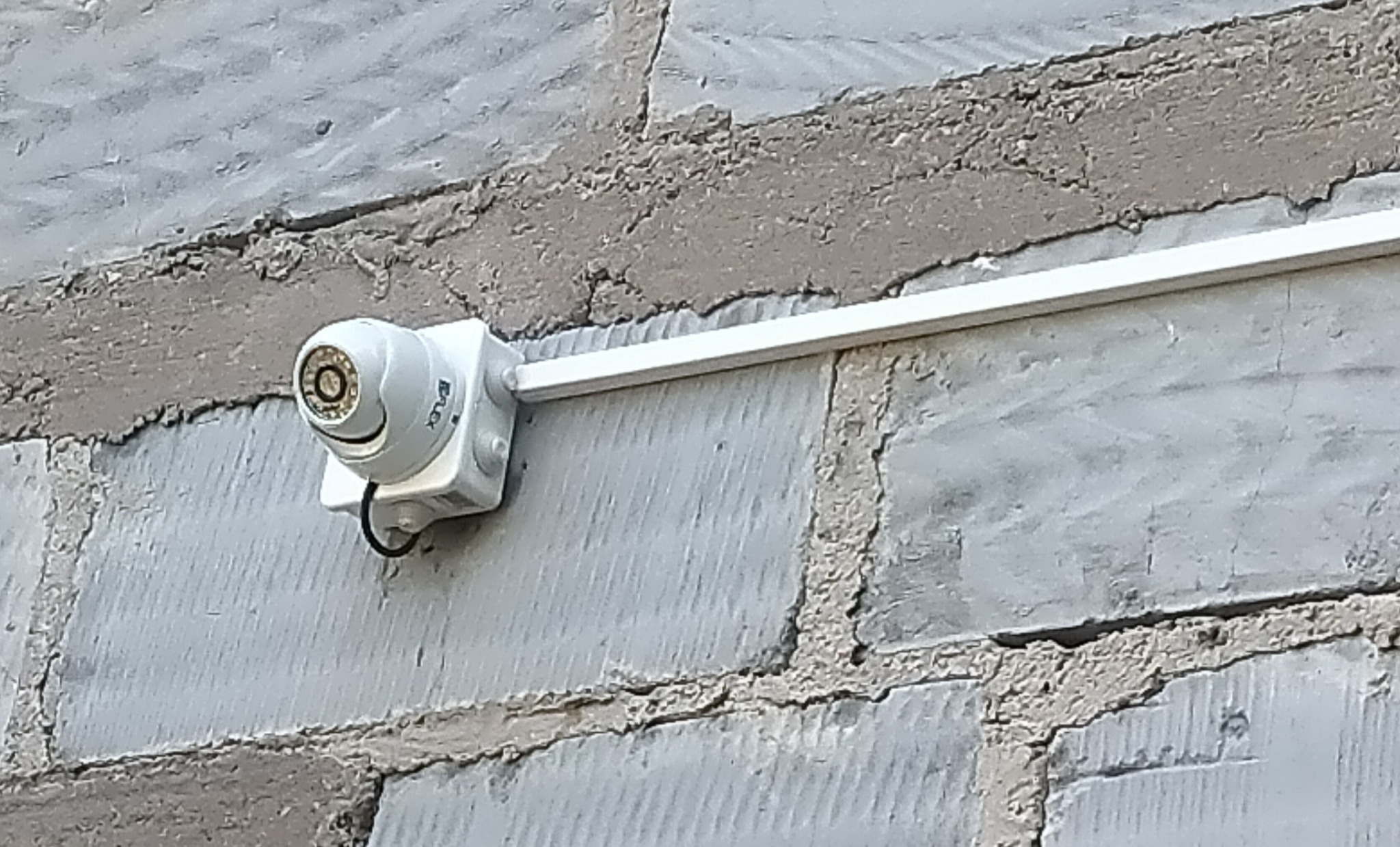 Kamera monitoringu IP zamontowana na szarej, surowej ścianie z cegieł. Widoczny kabel i biała listwa montażowa. Ujęcie z bliska, lekko z dołu.