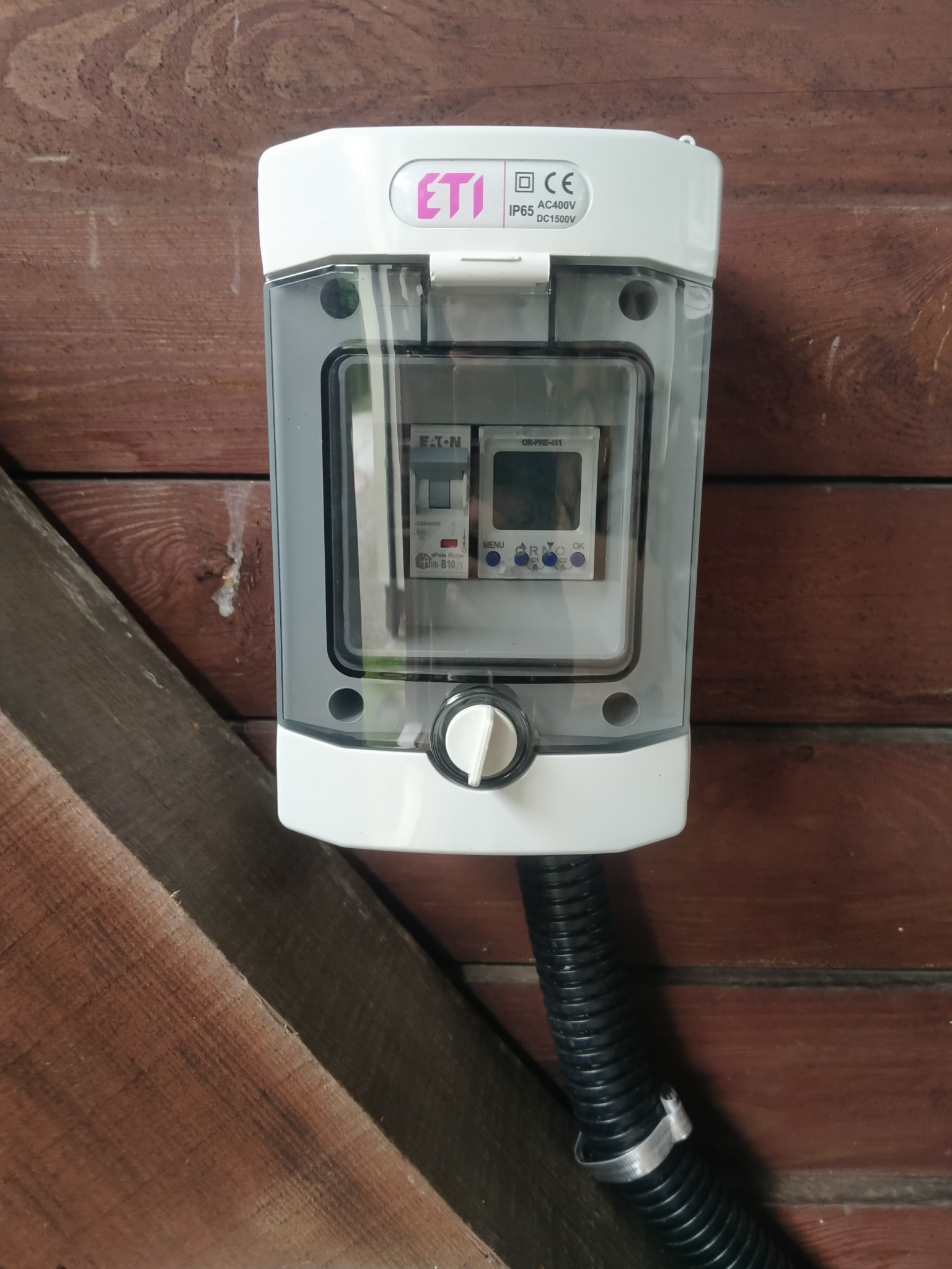 Szczegółowe ujęcie skrzynki elektrycznej ETI z zabezpieczeniami Eaton i programatorem ORNO, zamontowanej na drewnianej ścianie. Widoczny przewód w peszlu.