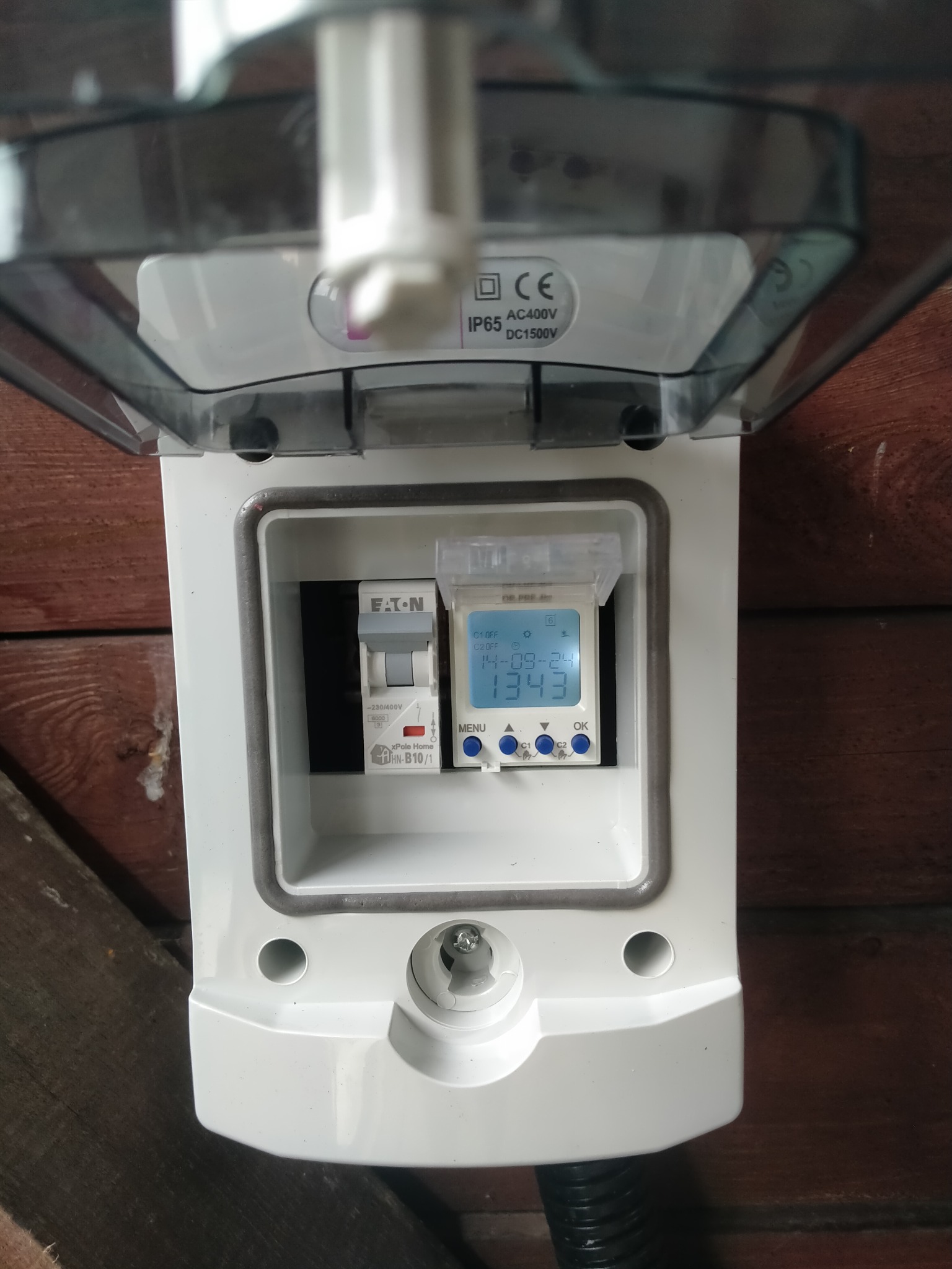 Biała skrzynka z otwartą pokrywą, wewnątrz wyłącznik Eaton i programator czasowy z wyświetlaczem LCD, zamontowana na drewnianej ścianie. Widoczne oznaczenia IP65 i CE.