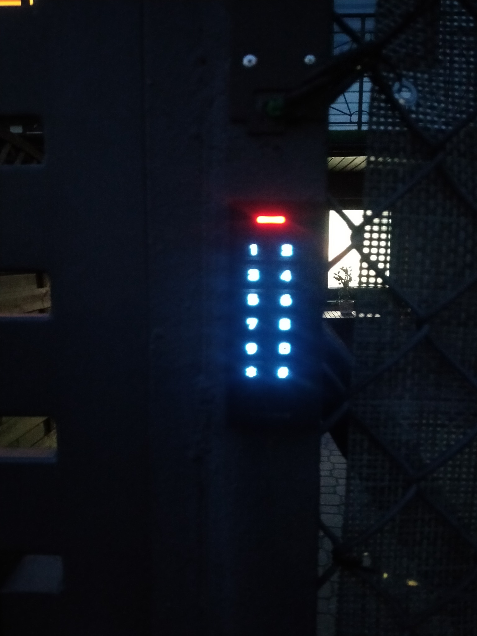 Podświetlona klawiatura numeryczna domofonu na tle metalowej bramy z kratą, widok z bliska, nocne oświetlenie.