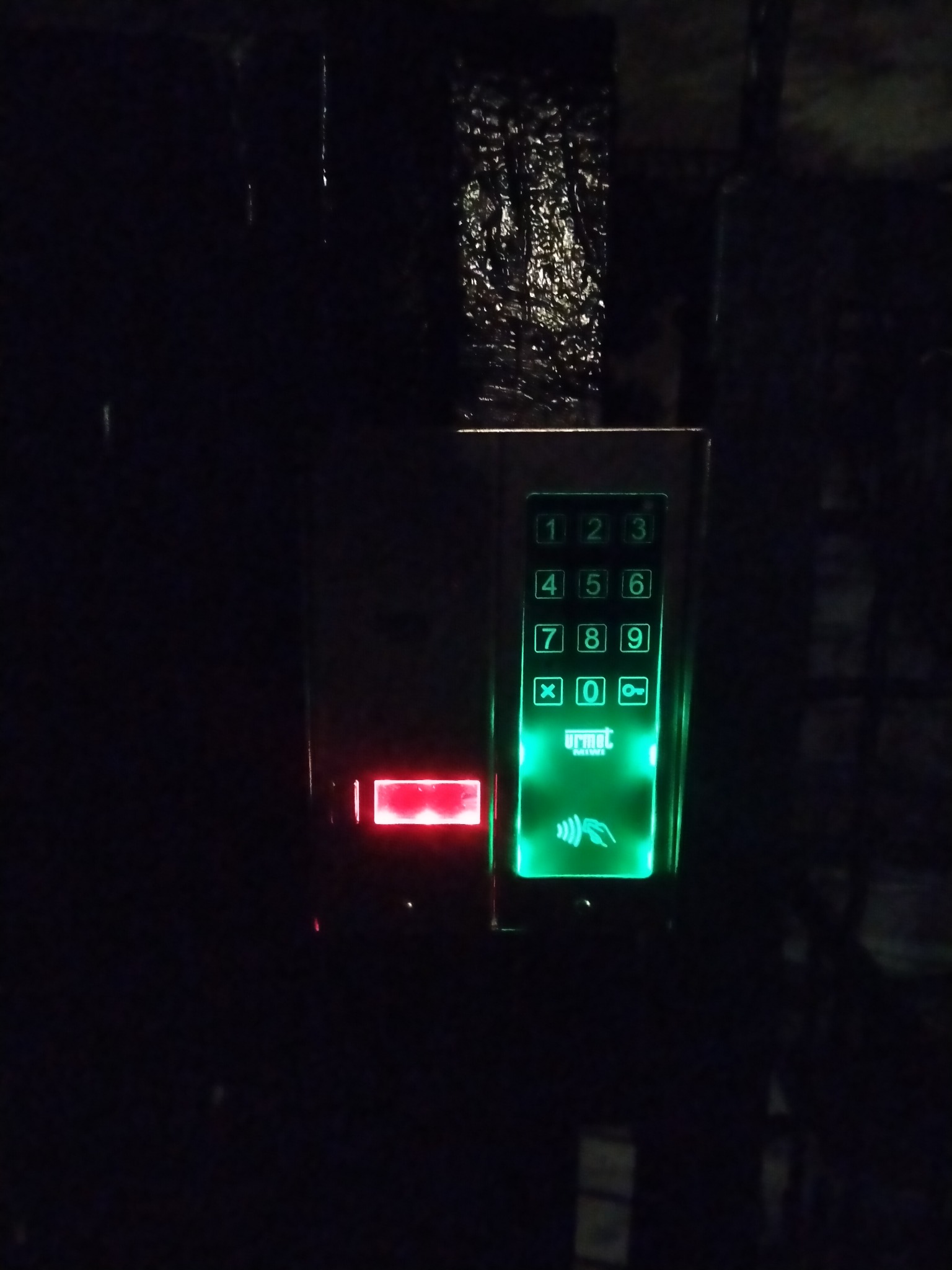 Podświetlony na zielono panel domofonu Urmet z klawiaturą numeryczną i czytnikiem kart zbliżeniowych, zamontowany na ciemnej ścianie. Widoczne czerwone podświetlenie.